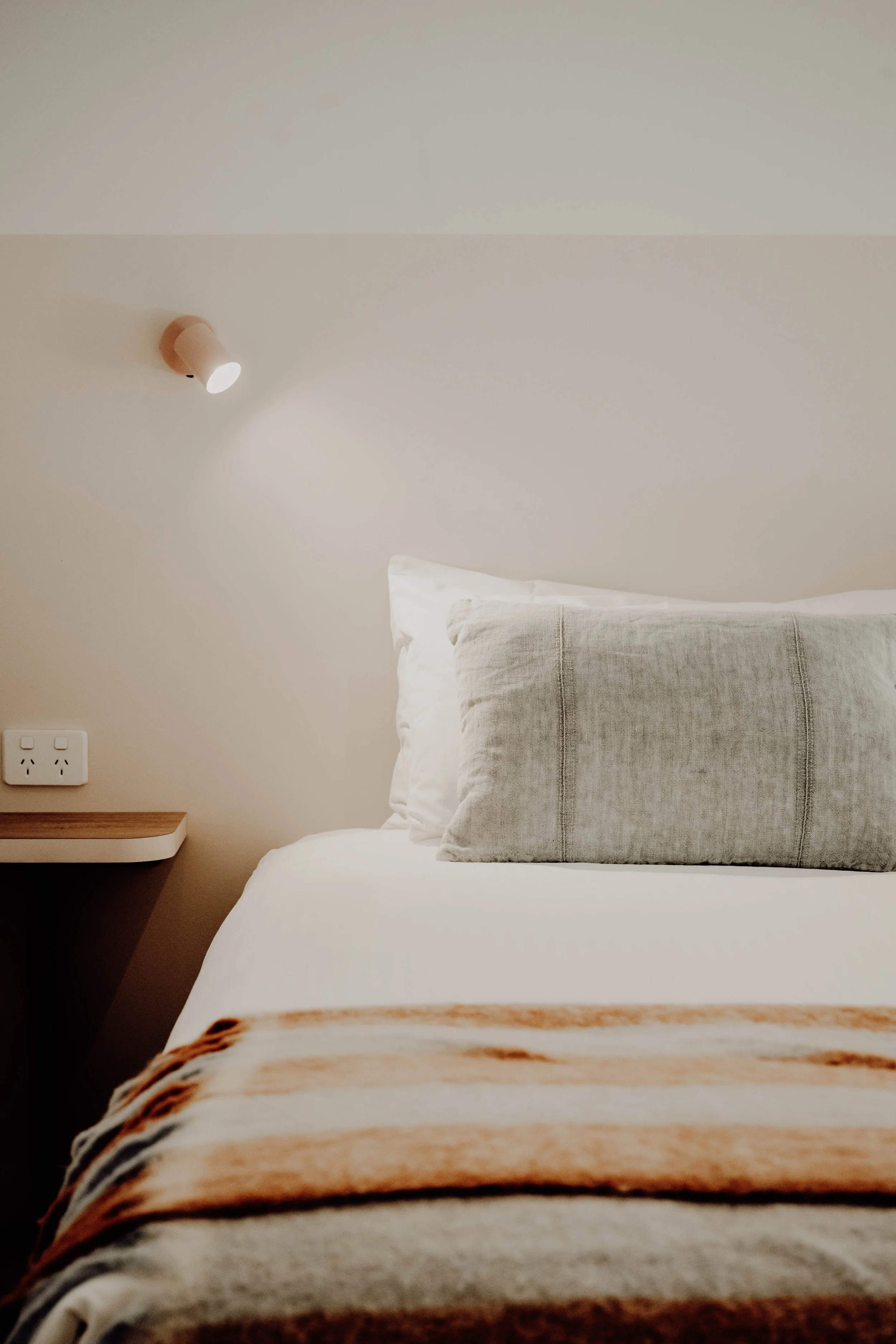 A neatly made bed with a gray pillow and white sheets in a minimalist bedroom. A wooden nightstand and a wall-mounted light fixture are visible.