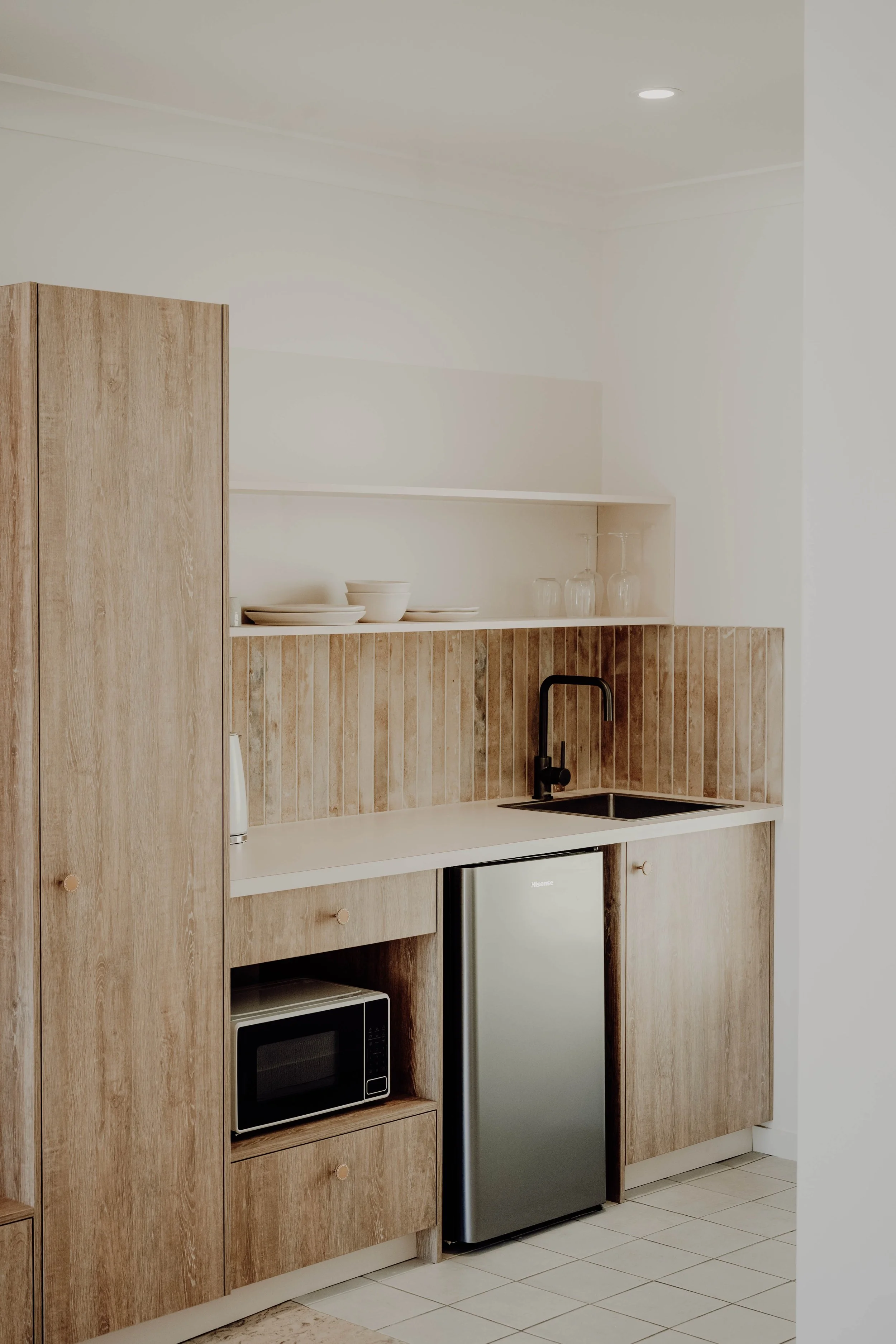 A compact kitchen with wooden cabinets, open shelves with dishes and glasses, a black faucet over a small sink, a mini fridge, and a microwave, featuring a beige tile backsplash.