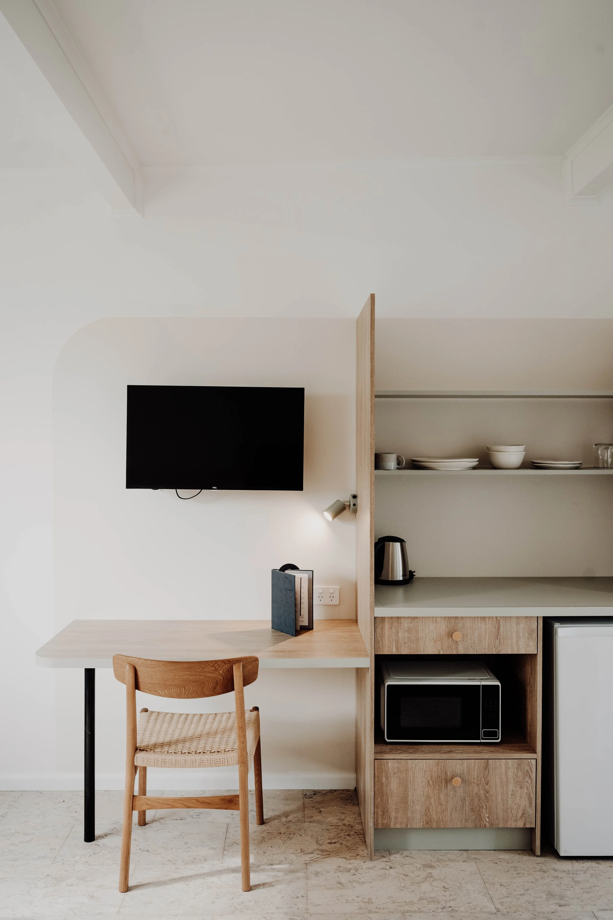 Interior of a minimalist kitchen corner with a wall-mounted TV, a small desk with a chair, open shelves with bowls and mugs, a kettle, a microwave, and a mini fridge.
