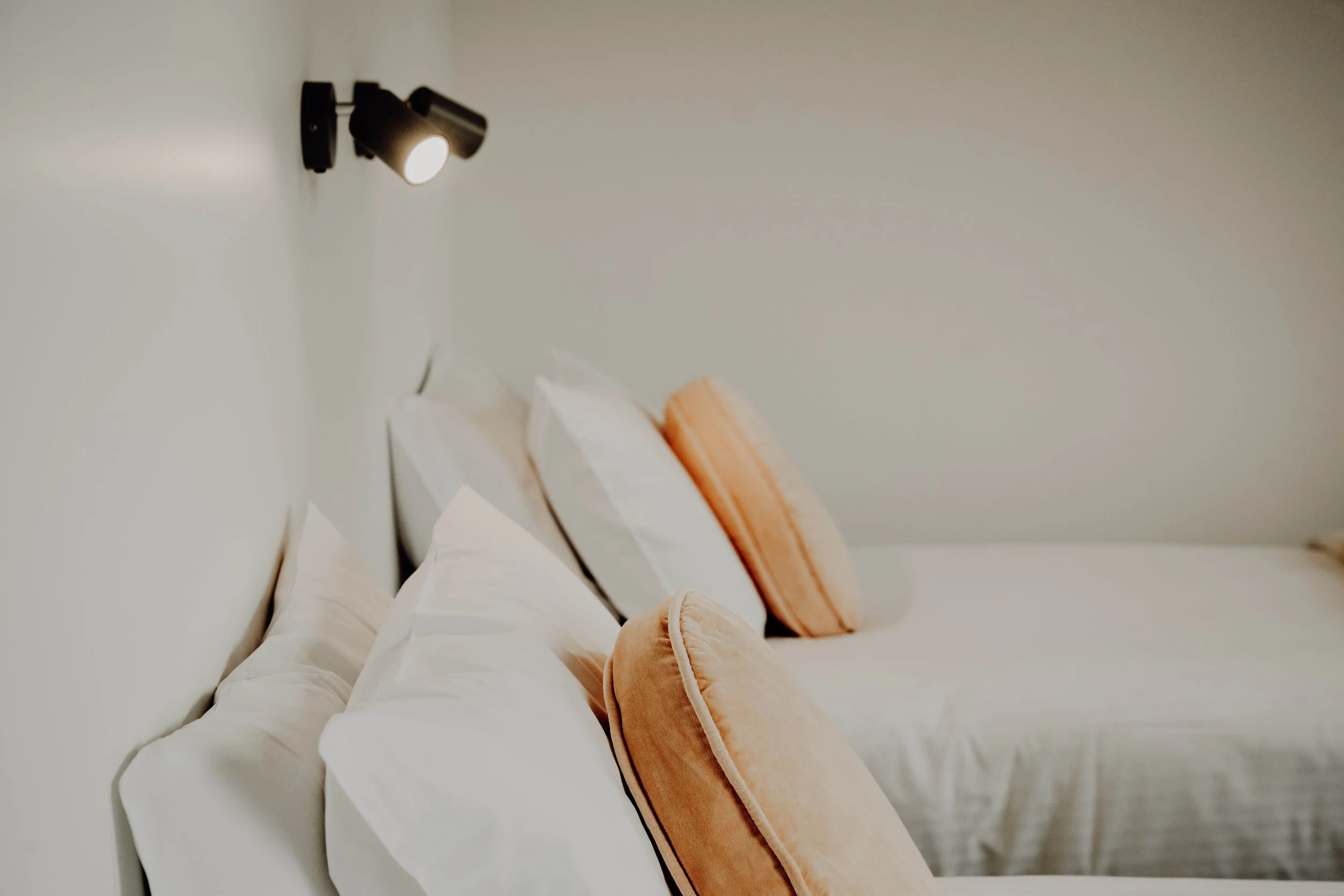 Two beds with white pillows and peach-colored bolster pillows, lit by a black wall-mounted reading lamp in a minimalist bedroom.