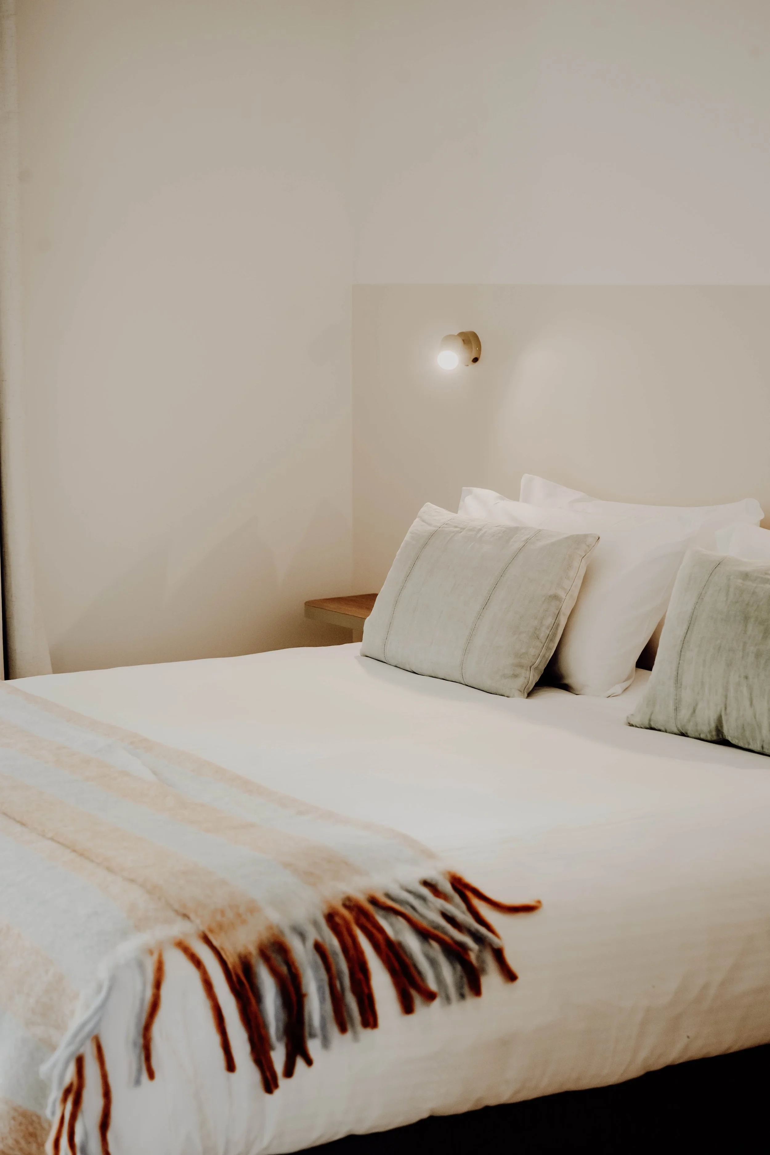 Made bed with white sheets, light-colored pillows, a beige blanket with fringed edges, and a wall-mounted light