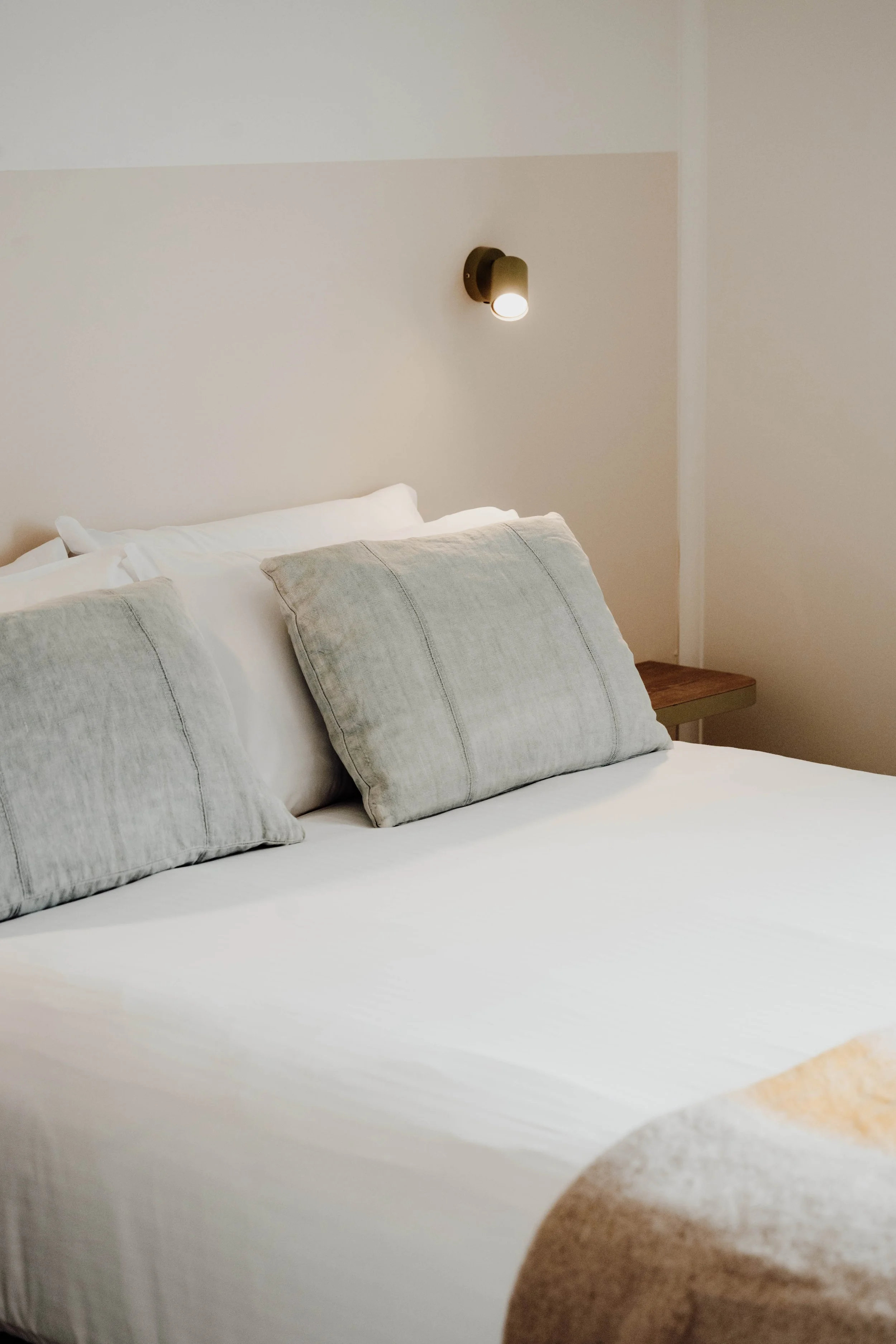 A neatly made bed with white sheets, gray pillows, and a beige blanket, in a modern bedroom with a wall-mounted light fixture.