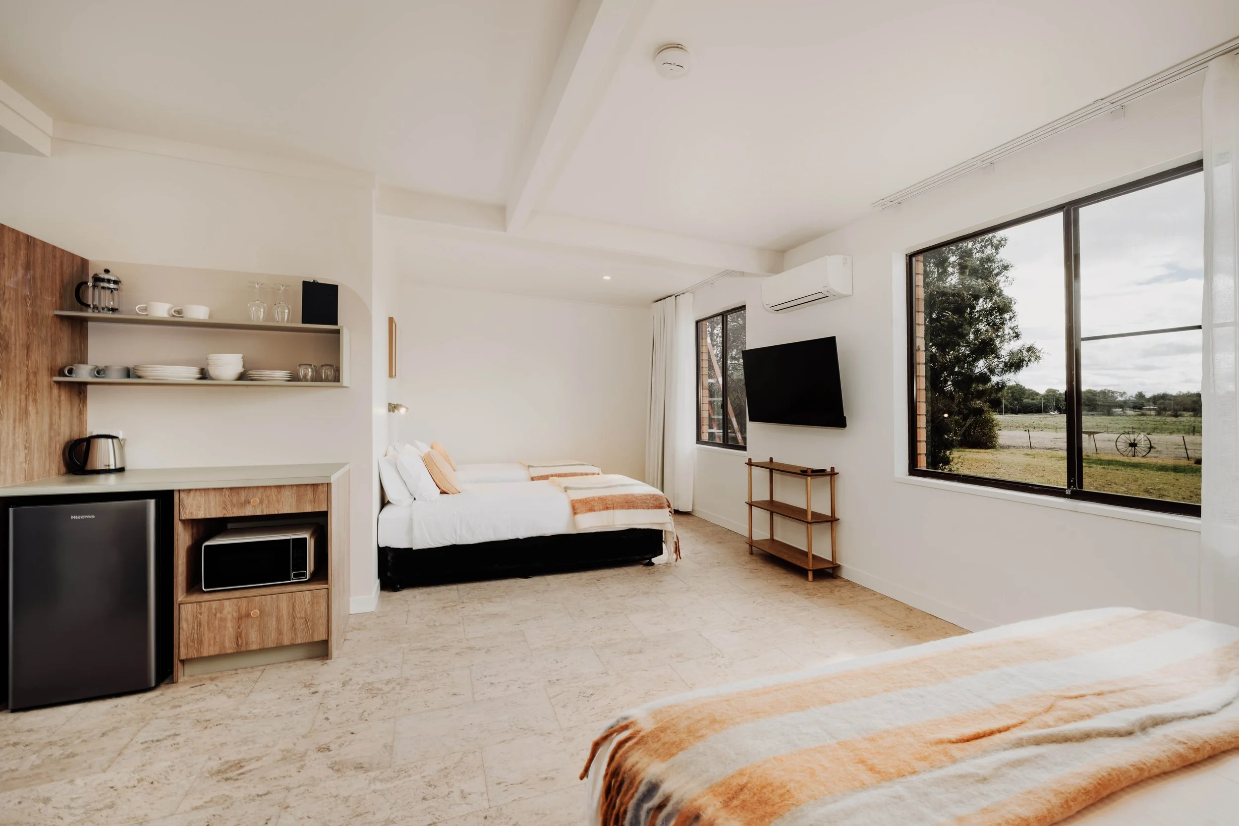 A bright hotel room with two beds, large windows showing greenery outside, a small kitchen area with a mini fridge, microwave, shelves with dishes, and a wall-mounted TV.