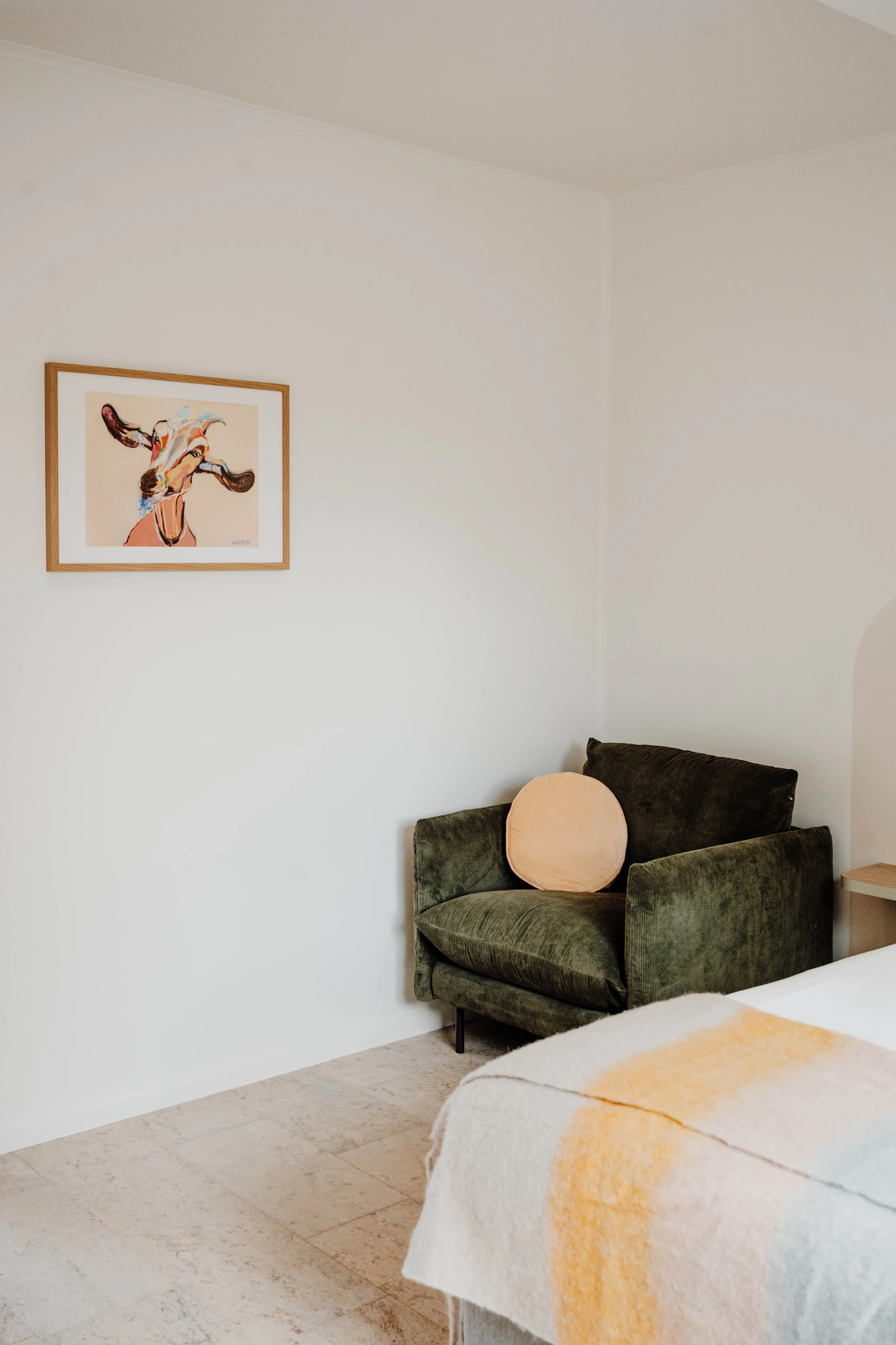 A corner of a room with a dark green velvet armchair, a round peach-colored pillow on it, a white bed with a peach and yellow blanket, light-colored flooring, a white wall with a framed painting of a cow, and a small wooden side table.