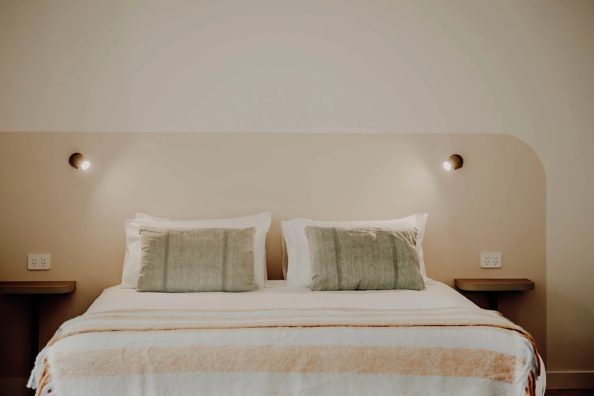 A neatly made bed with white and beige bedding, two pillows with greenish covers, and two wall-mounted reading lights on a minimalist bedroom wall.