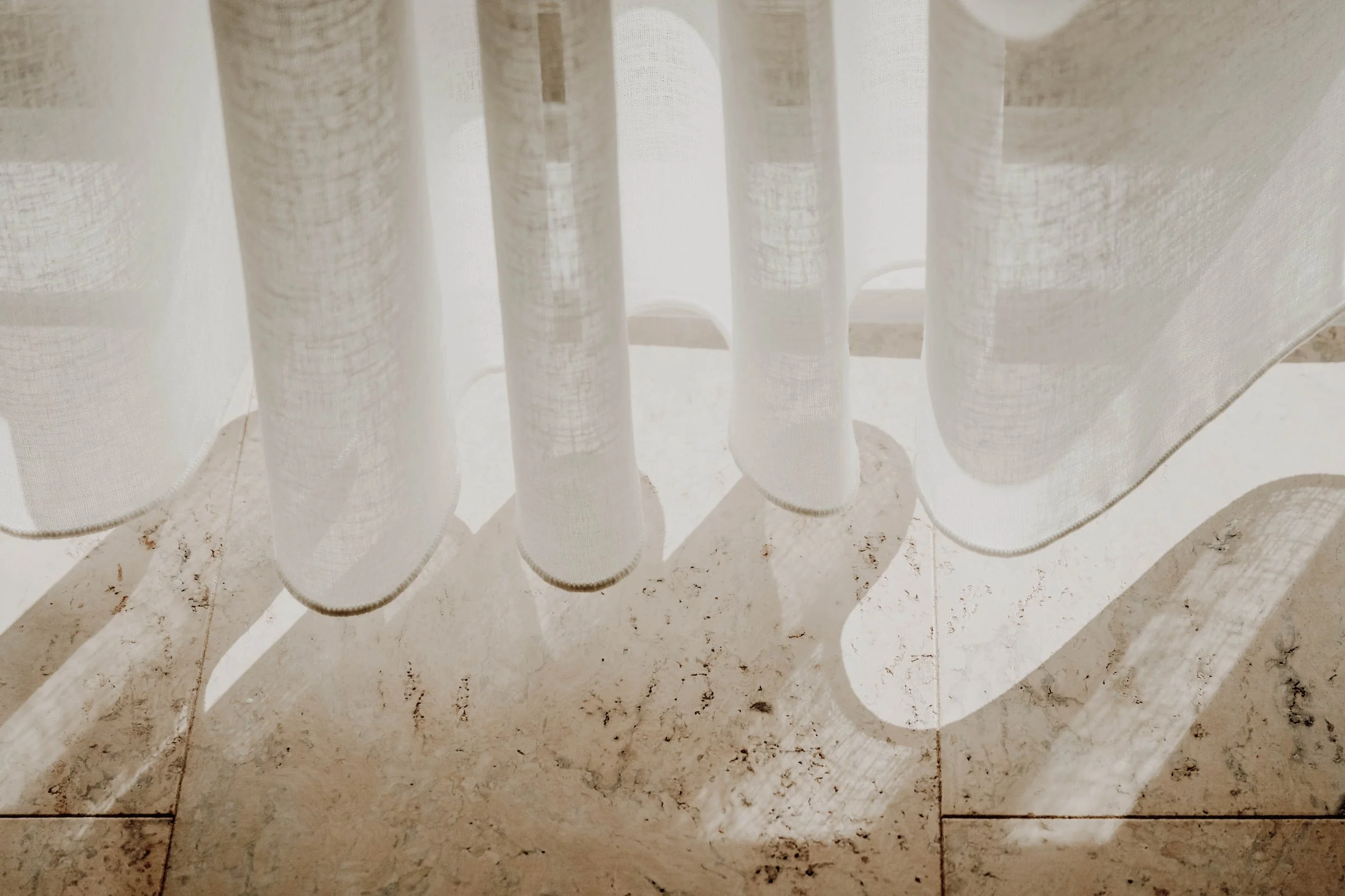 Close-up of sheer white curtains hanging in front of a bright window, casting shadow patterns on a cream-colored tiled floor.