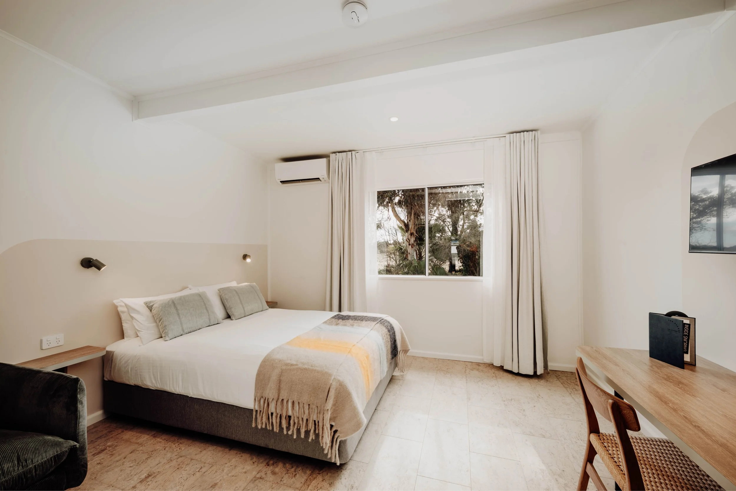 A bedroom with a large bed, light-colored bedding, and pillows, a window with white curtains showing trees outside, a wall-mounted TV, and a wooden desk with a chair.