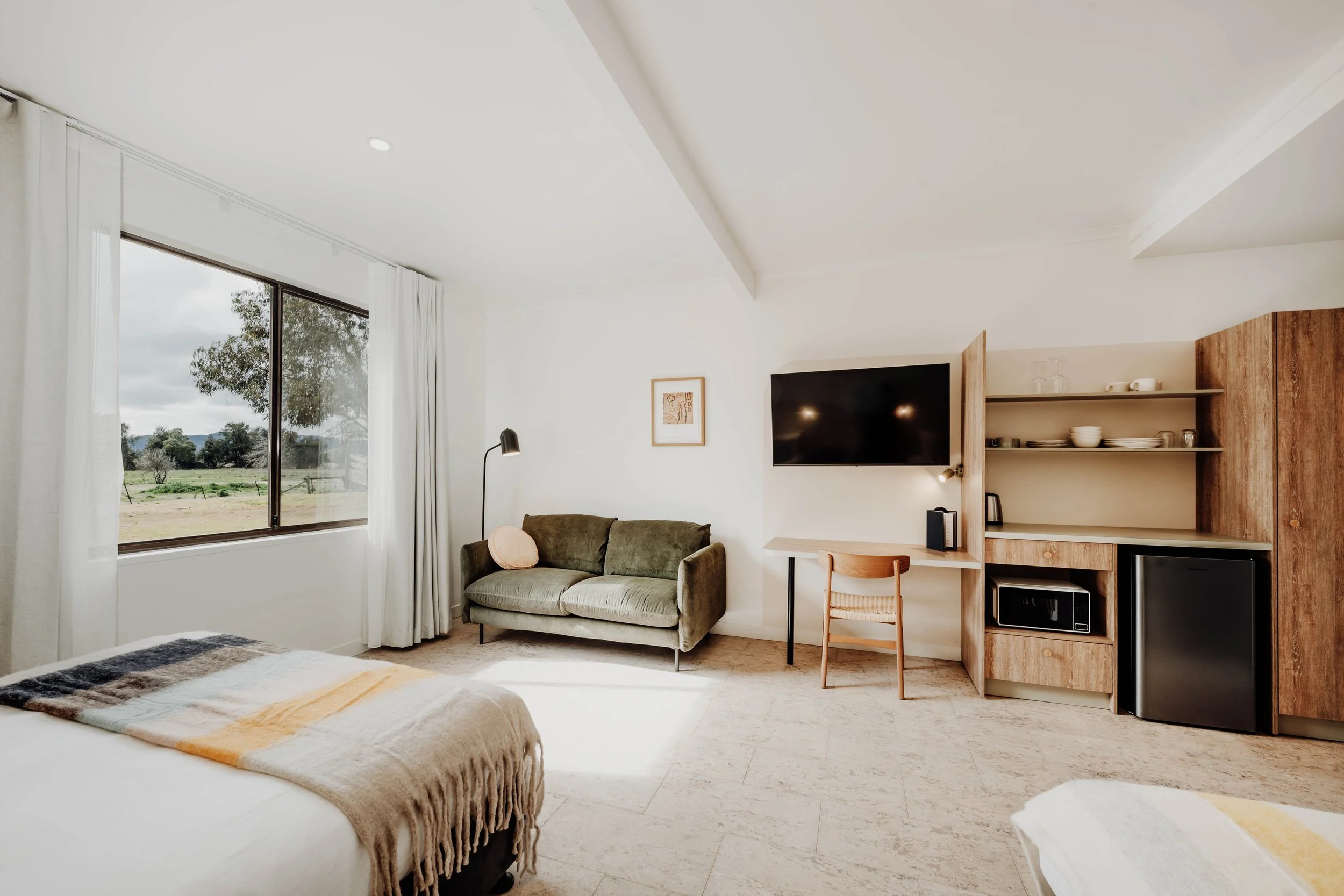 Bright hotel room with a large window showing an outdoor landscape, a green sofa with a pink pillow, a wall-mounted flat-screen TV, a small desk with a chair, and a wooden shelving unit with dishes, a microwave, and a mini fridge.