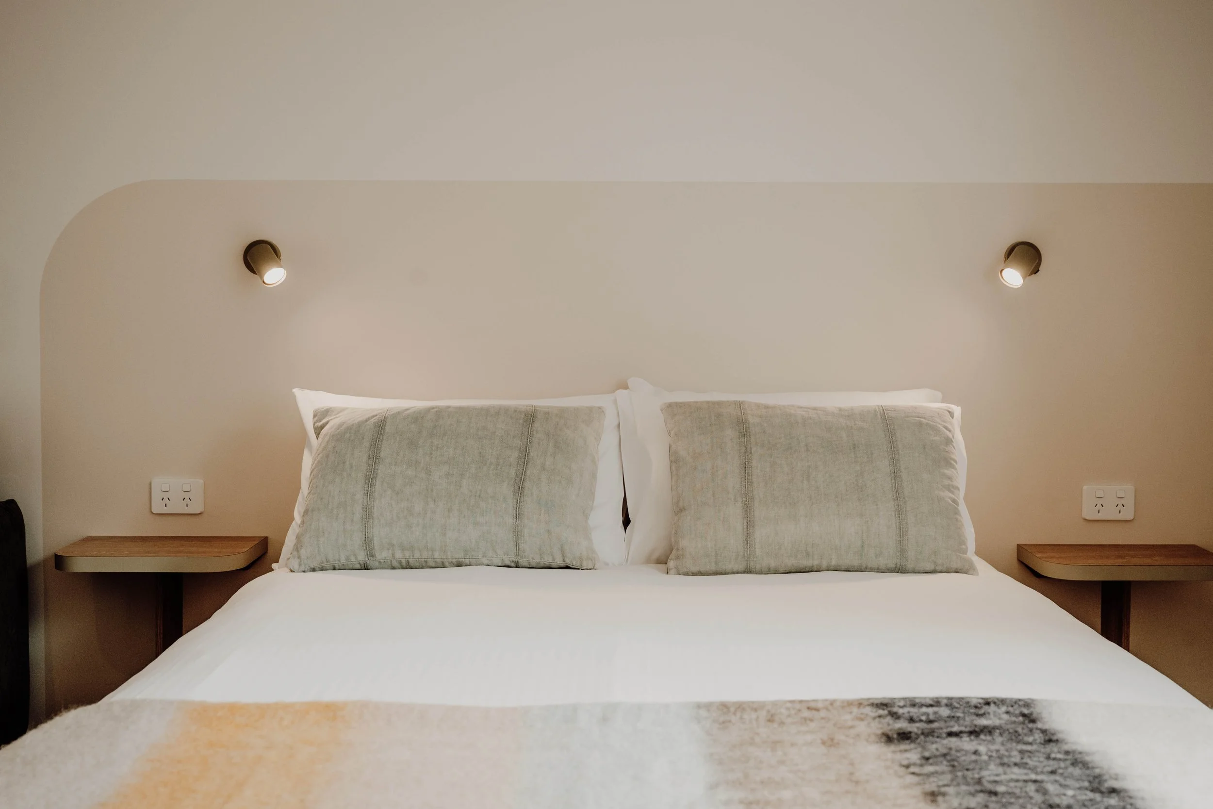 A neatly made bed with two large pillows and a beige headboard, with wall-mounted reading lights and small bedside tables on each side in a modern bedroom.