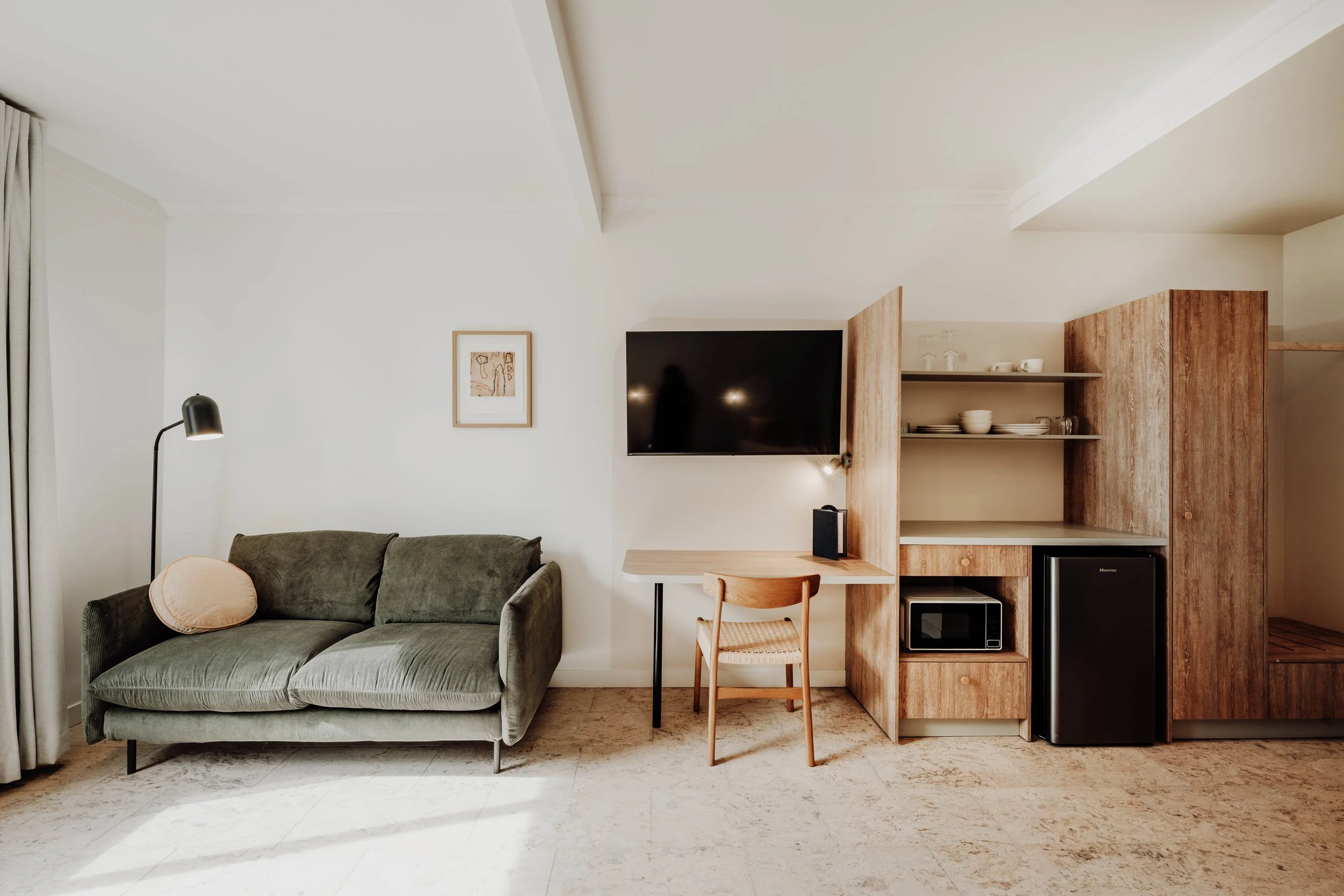 A living room with a dark green sofa, a reading lamp, and a wall-mounted TV above a small wooden desk with a chair. There is a wooden open shelving unit with dishes and a microwave, and a mini refrigerator underneath.