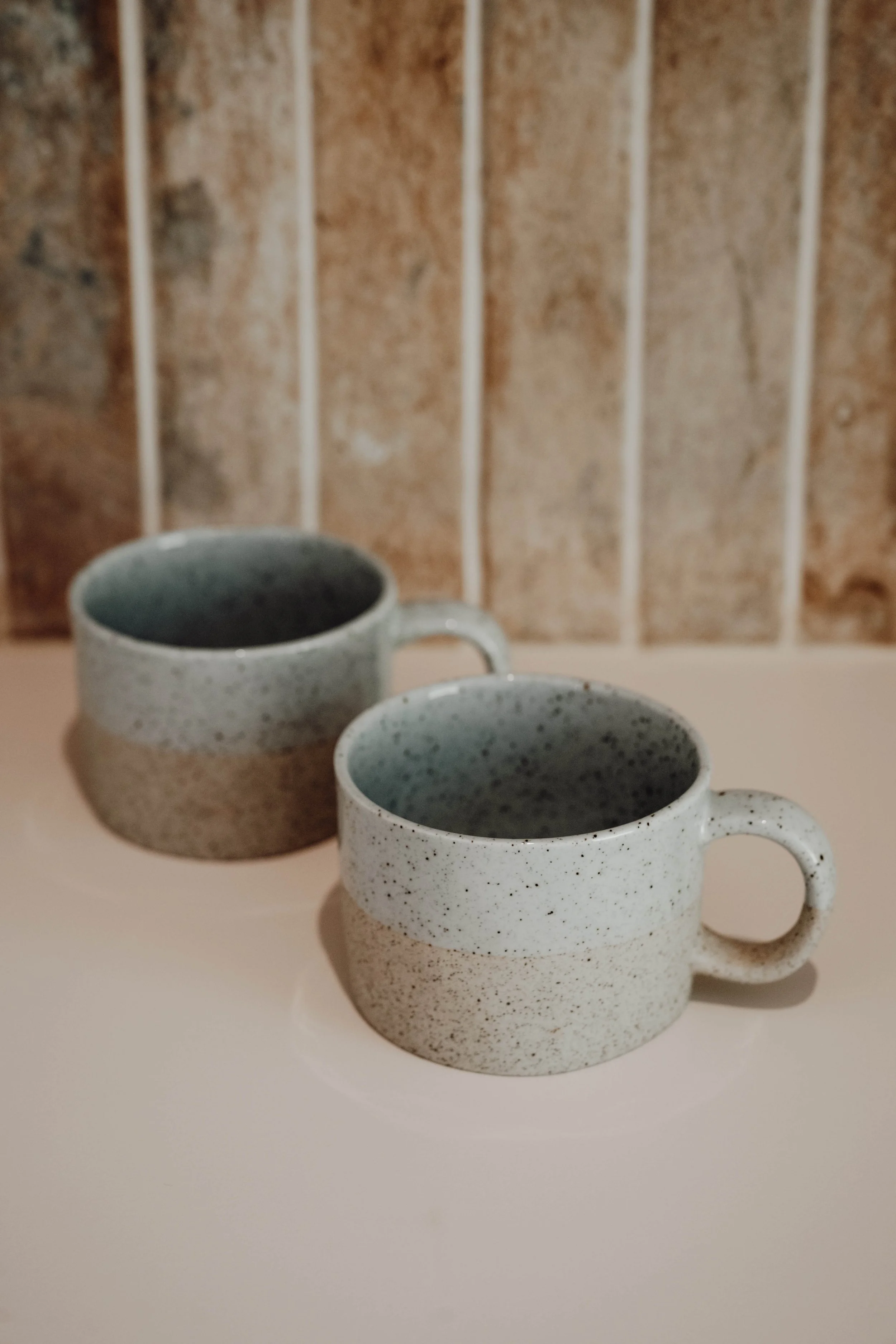 Two speckled ceramic coffee mugs placed on a light-colored surface with a brown textured wall in the background.