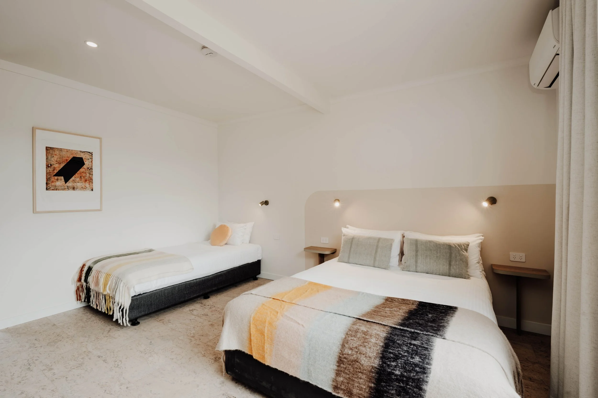 A bedroom with a large bed with beige and white bedding, two pillows, and a wall-mounted reading light. To the left, there is a small single bed with white bedding, a peach pillow, and a framed artwork on the wall. The room features neutral tones, an