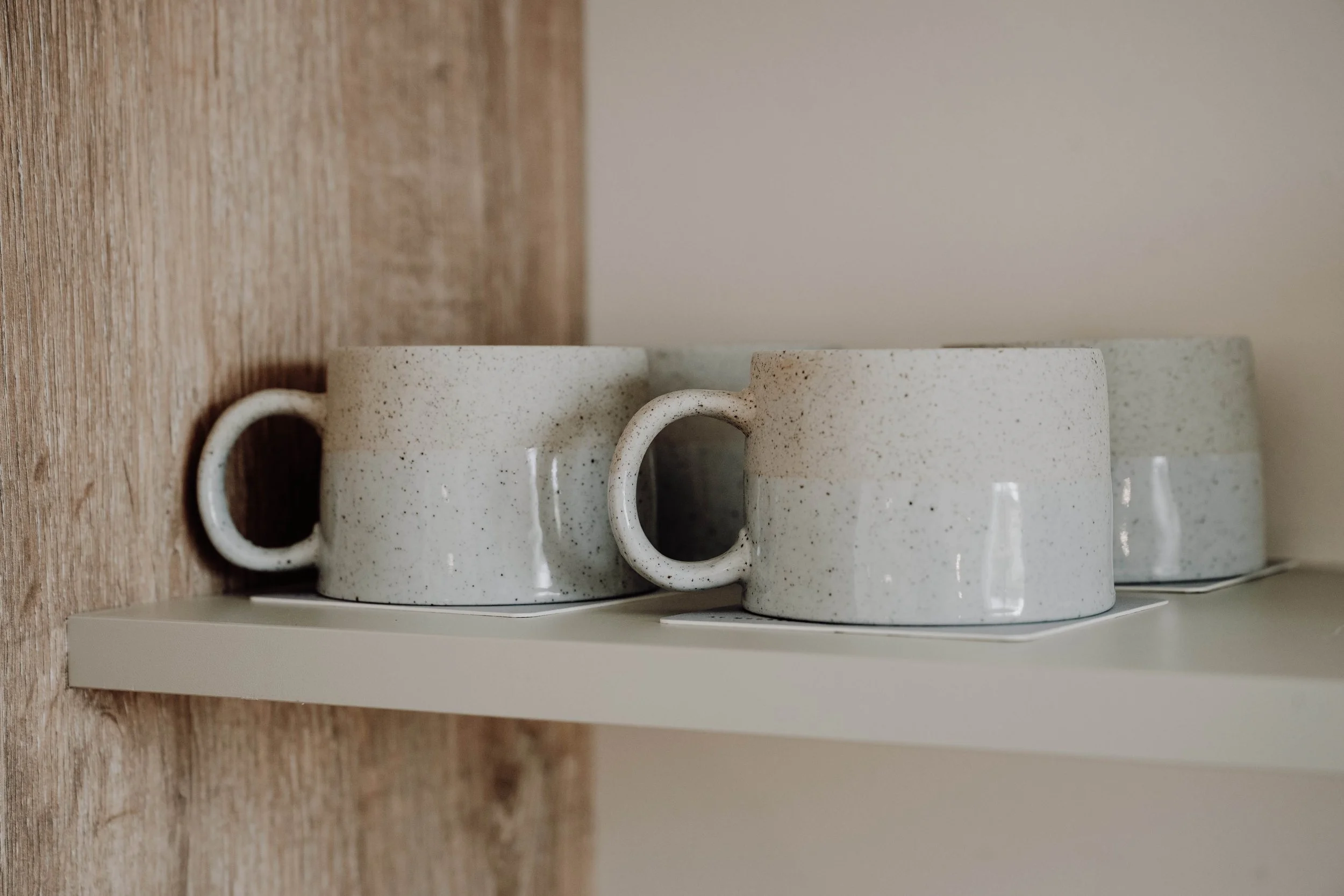 Three speckled ceramic cups placed upside down on a white shelf, with a wooden wall to the left and a white wall in the background.
