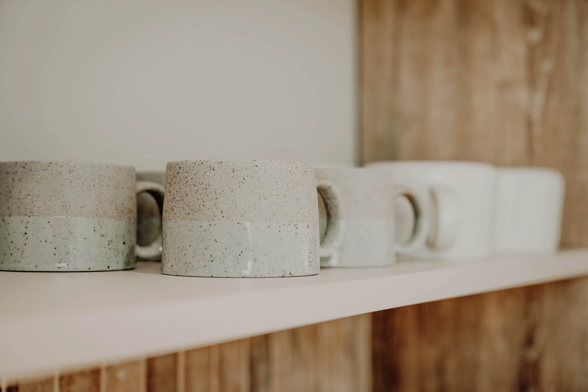 Cream-colored speckled ceramic mugs on a white shelf against a wooden wall.