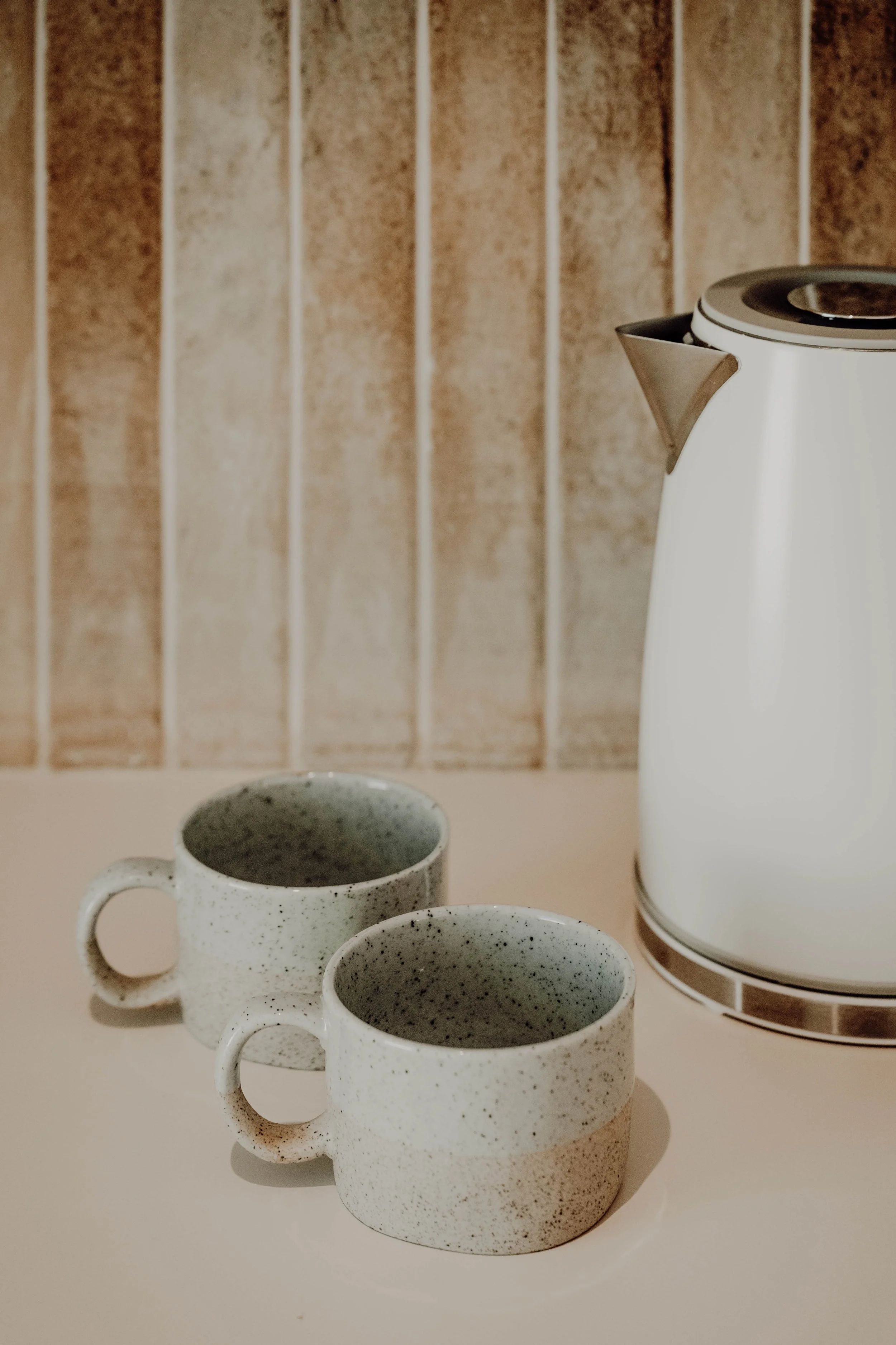 Two speckled ceramic coffee cups and a white electric kettle on a light-colored surface with a wooden panel background.
