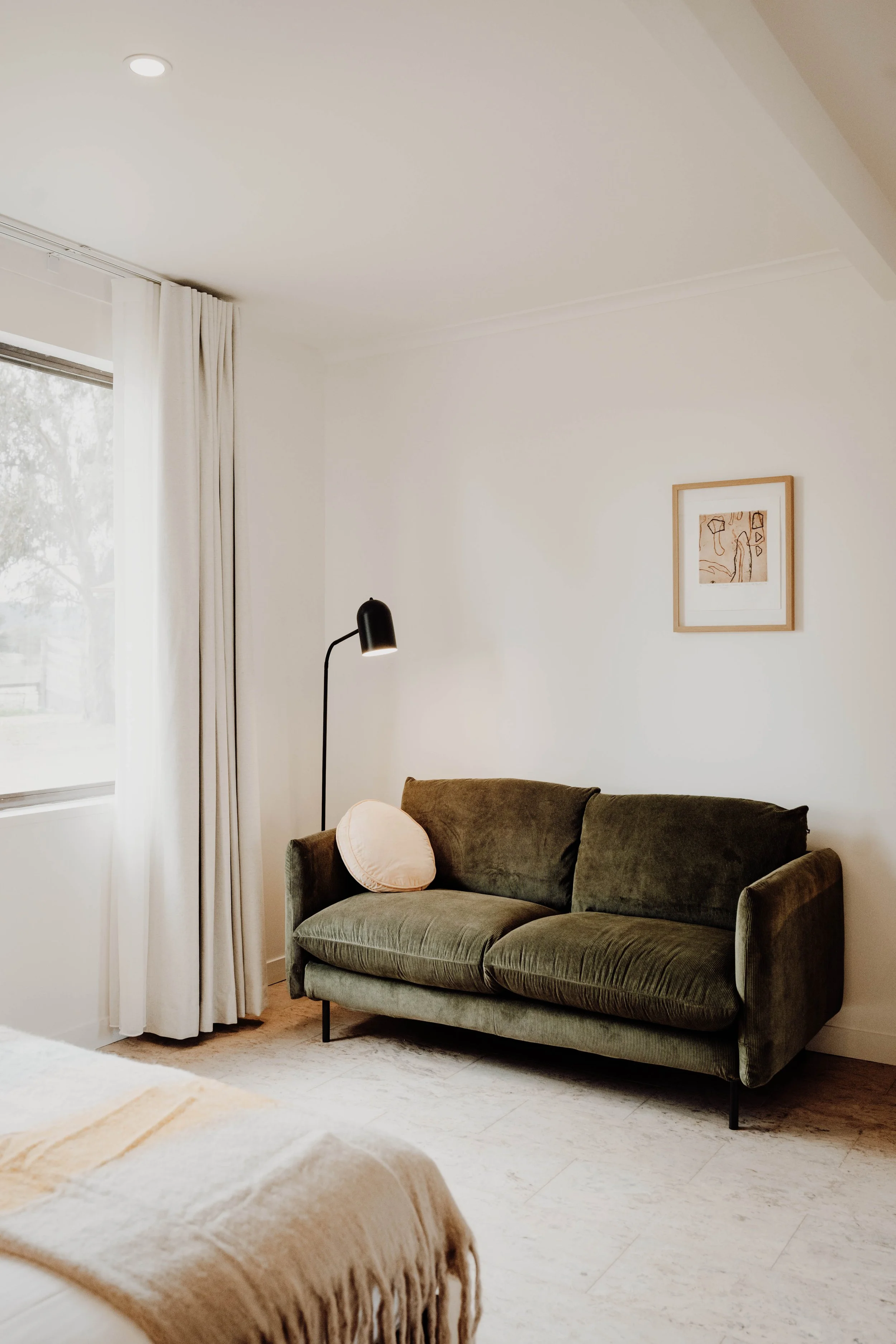 A cozy living room corner with a green sofa, a pink pillow, a black floor lamp, a window with white curtains, and a framed artwork on a white wall.