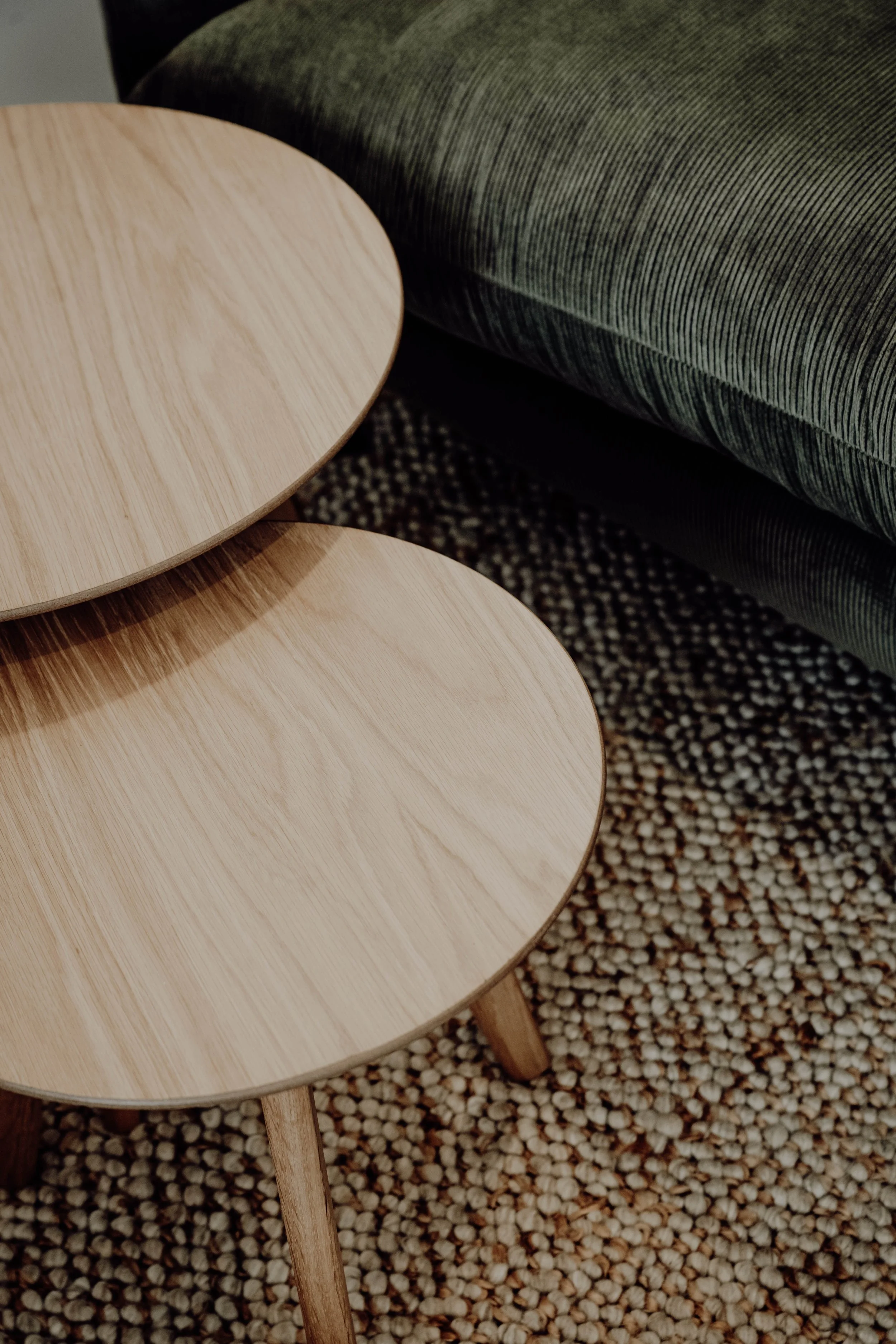 Close-up of two round wooden coffee tables with thin legs, situated on a patterned carpet, next to a green upholstered sofa.