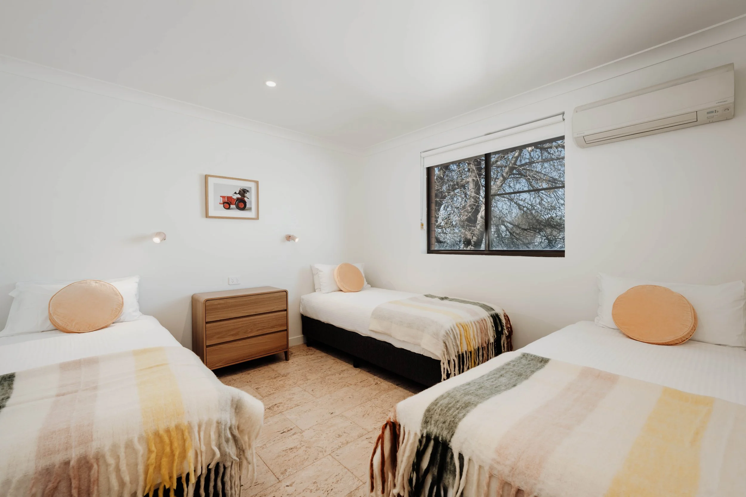 A bedroom with three single beds, each with white bedding, round orange pillows, and multicolored striped blankets. There is a wooden dresser between the beds, a window with a view of tree branches outside, and an air conditioning unit on the wall. A