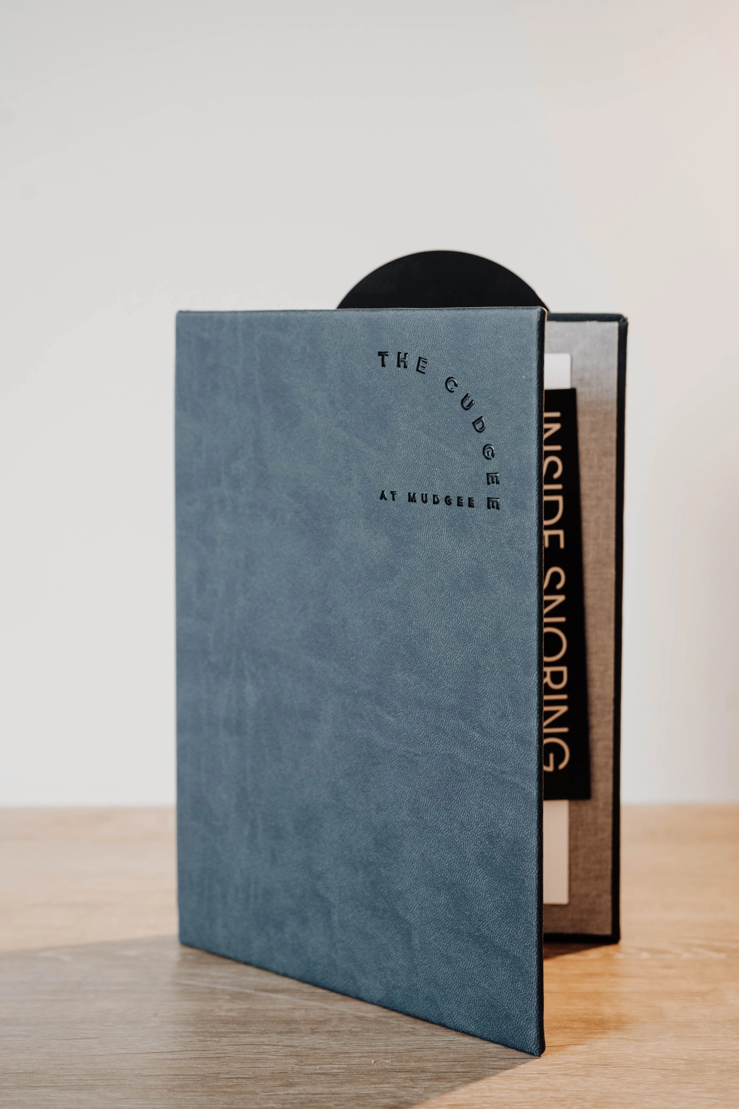 A blue menu titled 'The Guide at Mudgee' standing upright on a light-colored wooden table, partially covering a black and beige menu.
