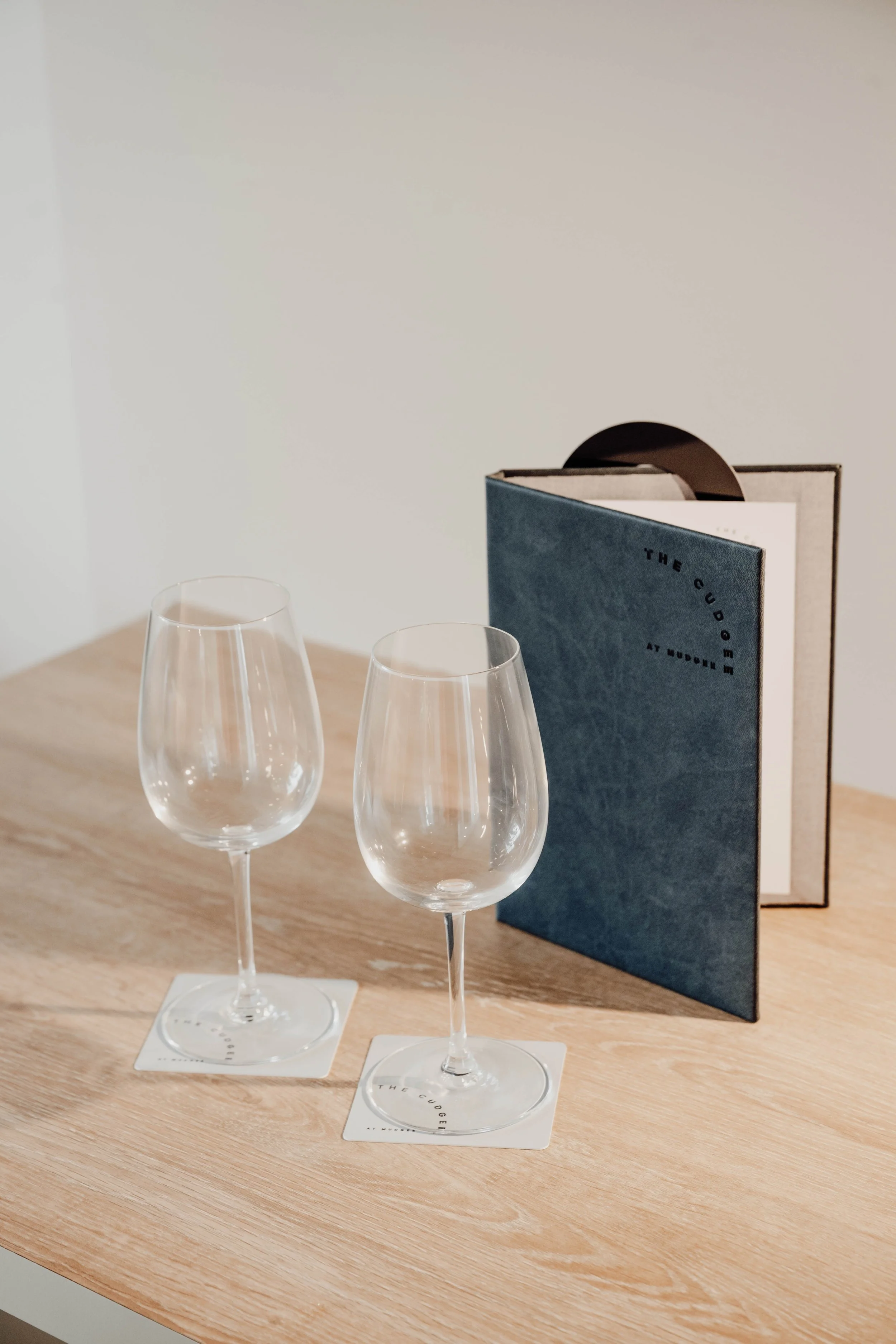Two empty wine glasses on coasters are placed on a wooden table. Behind them is a blue menu and a brown menu standing upright.