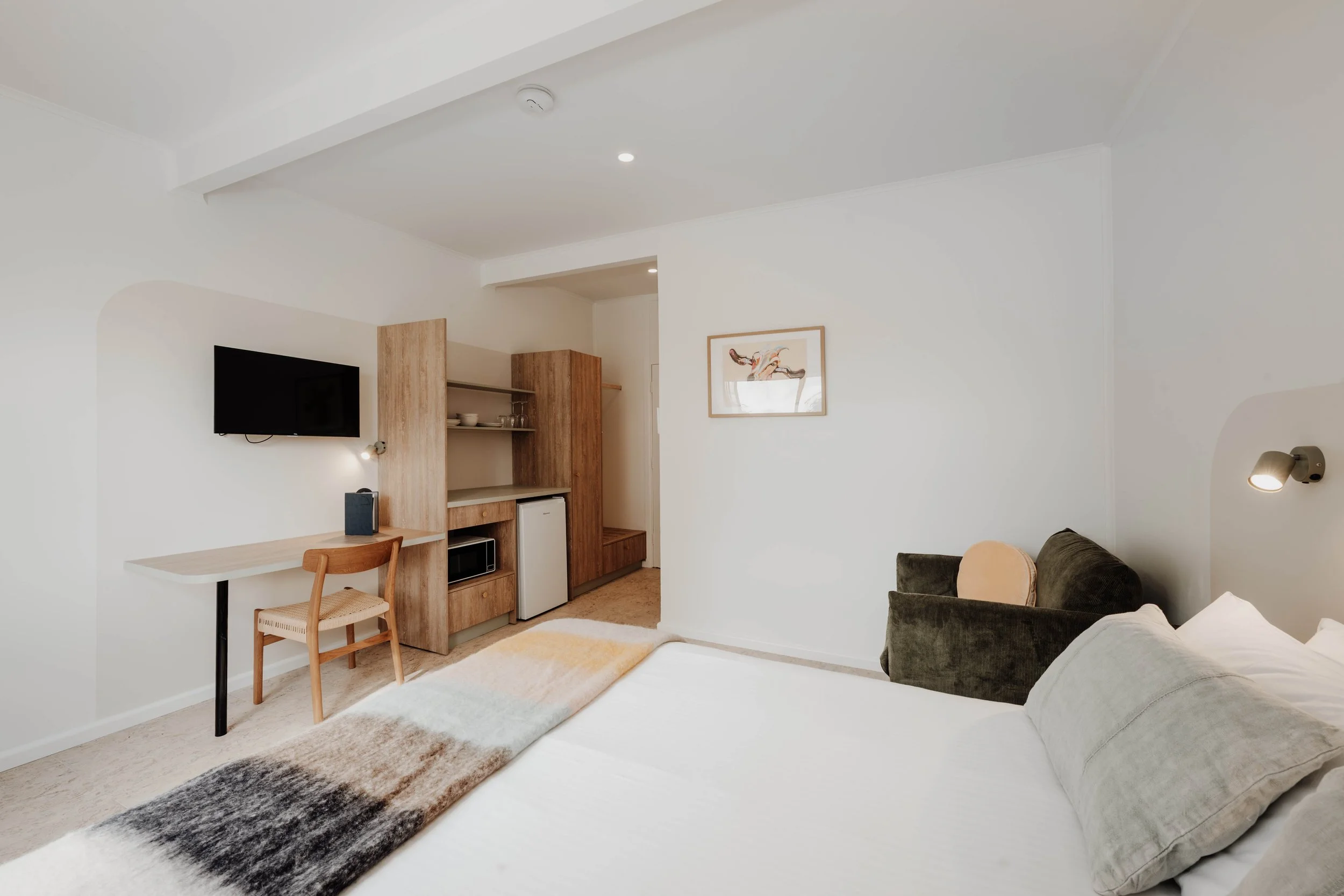 Modern hotel room with a bed, green armchair, wall-mounted TV, wooden desk, and minimal decor.