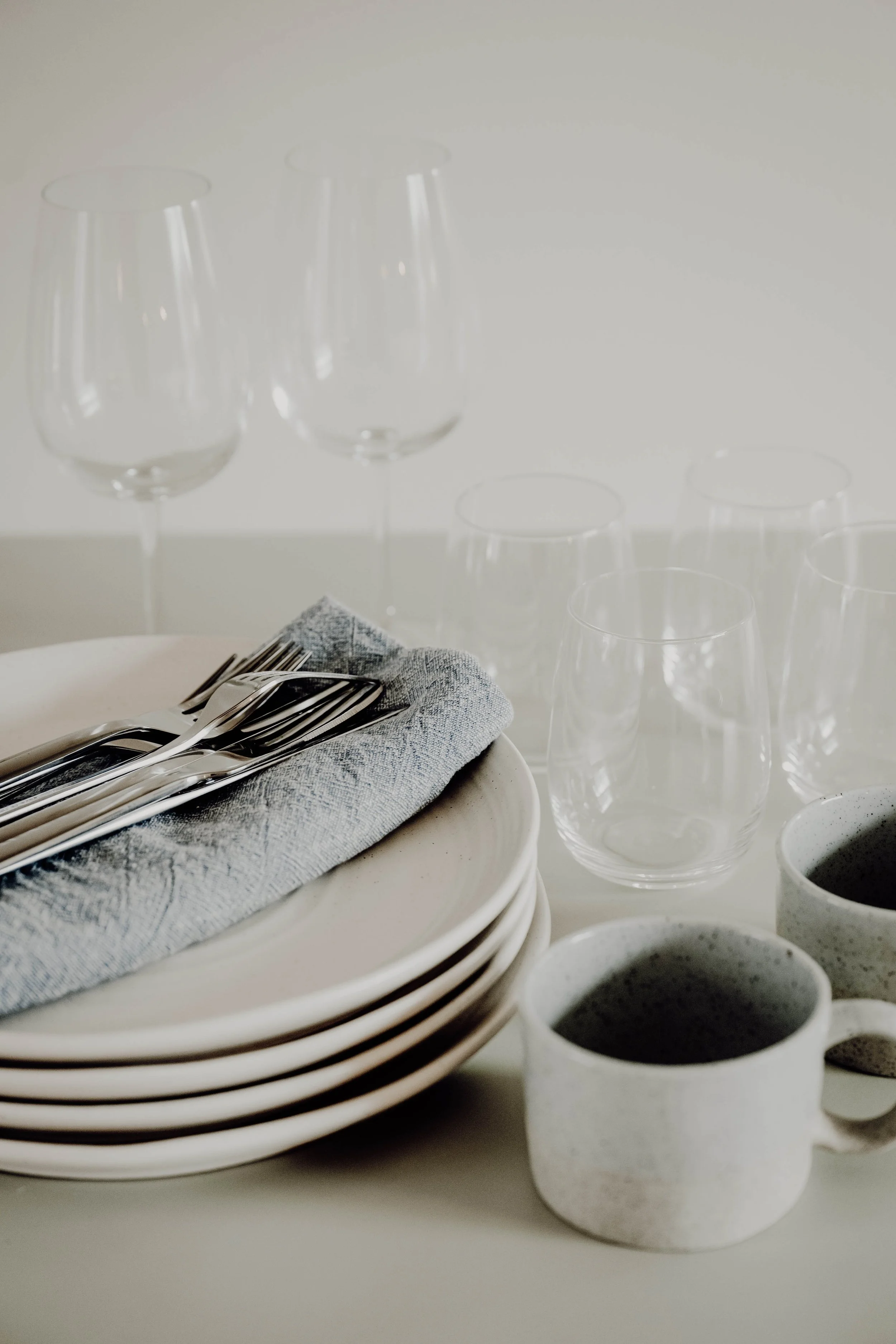 Stack of white plates with a gray napkin and silverware on top, surrounded by empty wine glasses and coffee mugs on a light table