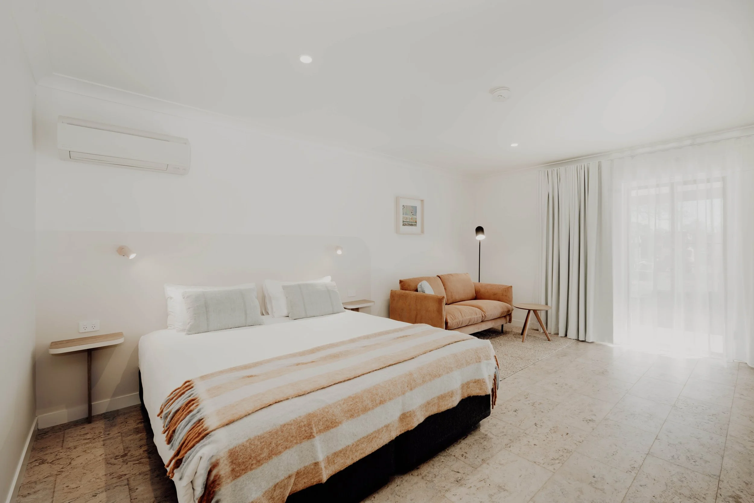 A spacious hotel room with a large bed, a small bedside table, a cozy orange sofa, a floor lamp, and a small side table near a large window with sheer curtains, bright and minimal decor.