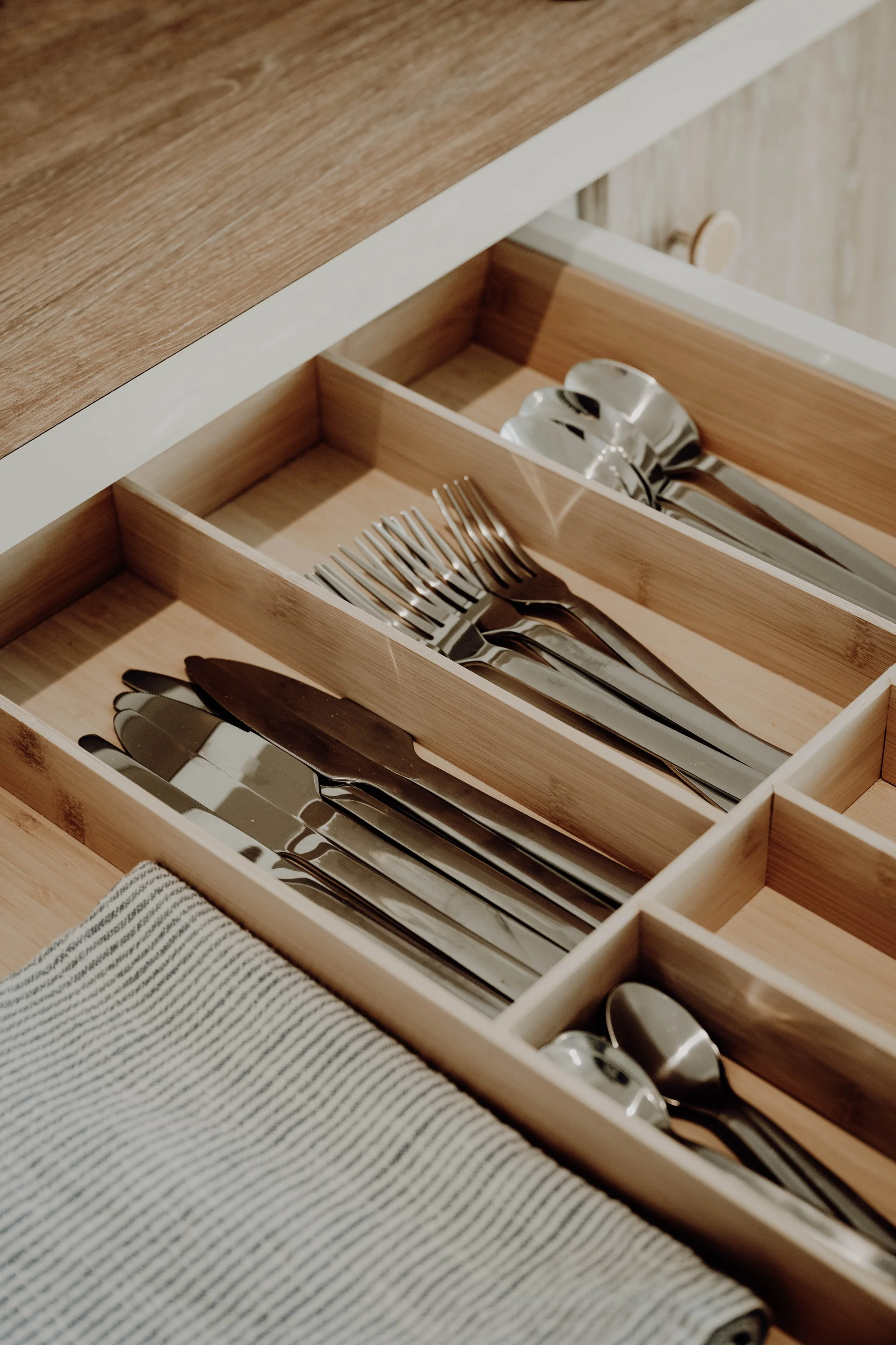 Drawer containing neatly arranged stainless steel forks, knives, and spoons, with a black and white striped cloth partially visible.