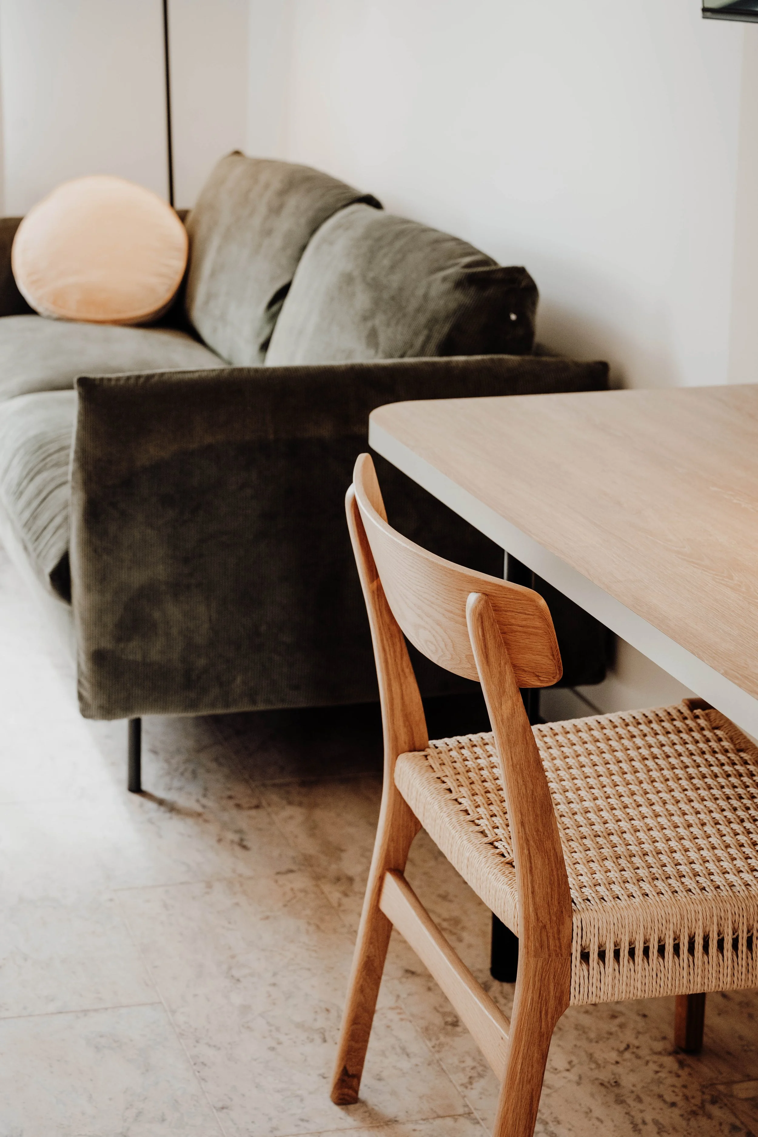 A view of a living room space showing a gray couch with a peach-colored cushion on it, a wooden dining chair with woven seat, and a wooden dining table.