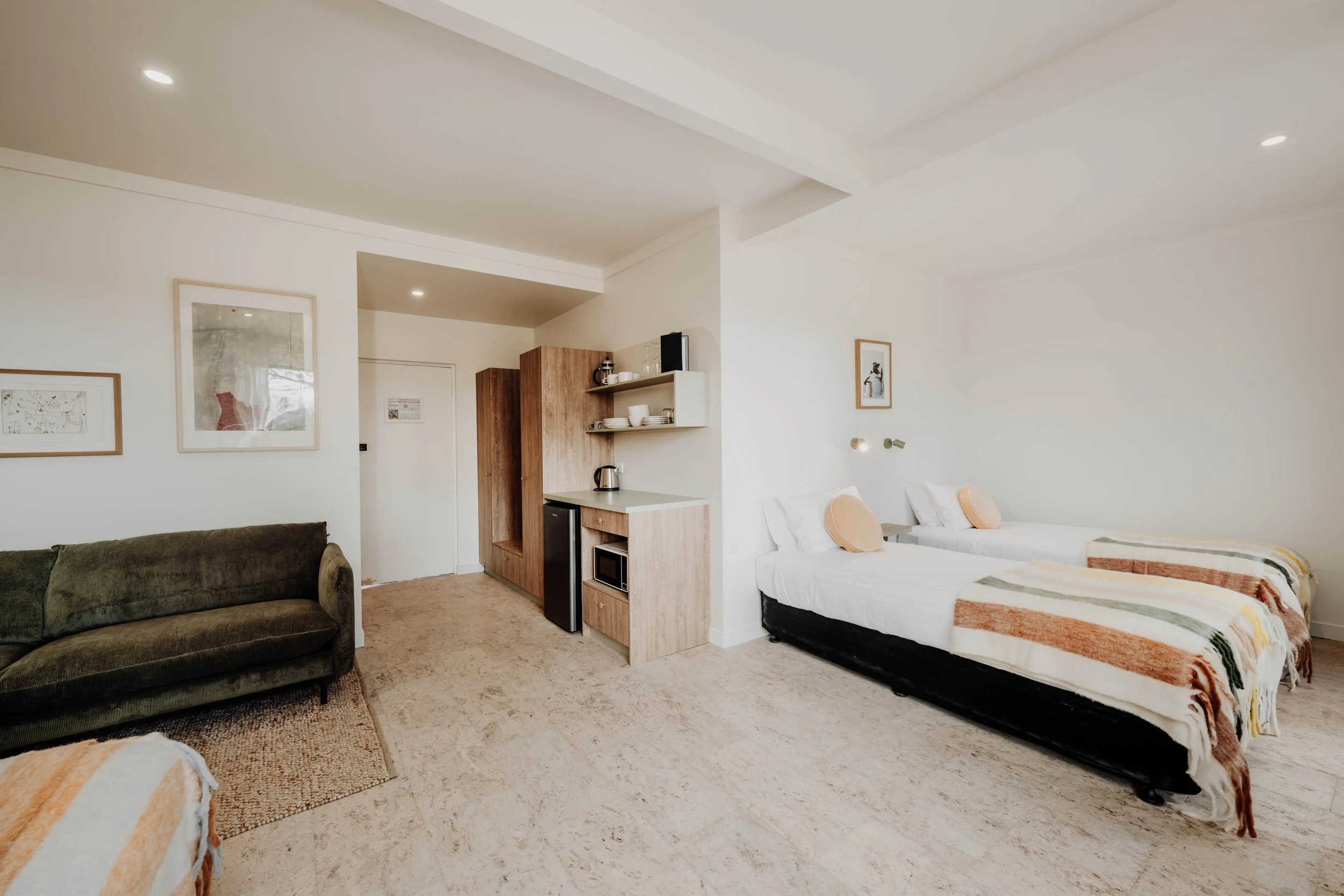 A hotel room with two beds, a green sofa, a small kitchenette, and framed artwork on the walls in a modern, minimalist design.