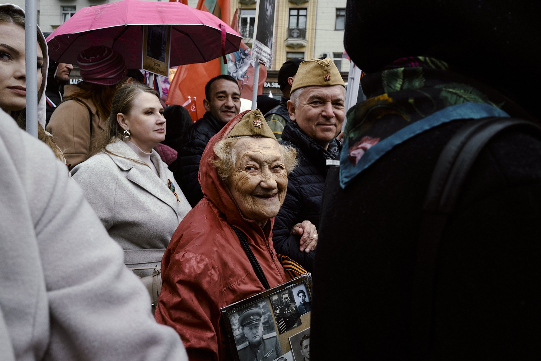 Eine Gruppe von Menschen bei einer Veranstaltung, darunter eine ältere Frau mit einem Fotoalbum von Soldaten, einige tragen Mützen mit Rotstern, vermutlich beim Gedenken an den Zweiten Weltkrieg.