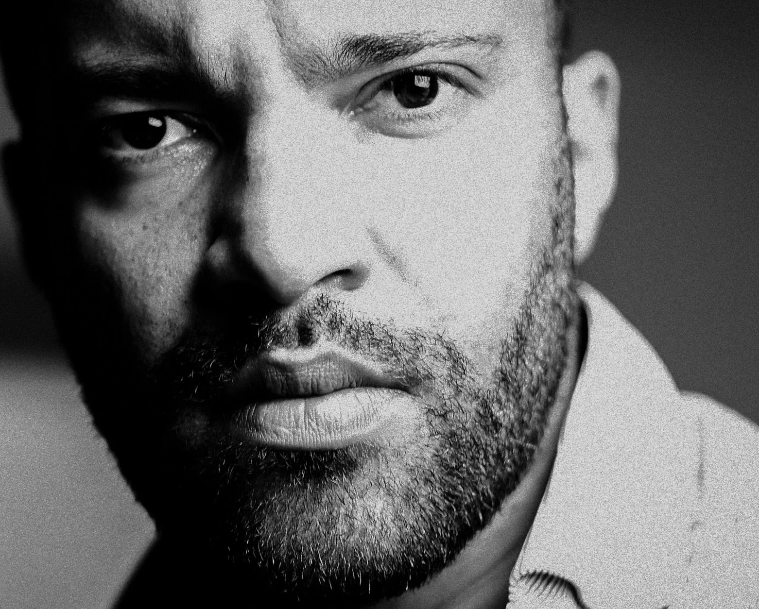 Schwarz-Weiß-Fotografie eines Mannes mit expressive Gesichtsausdruck, dichter Bart, leicht gefurchter Stirn und kurzem Haar, blickt direkt in die Kamera.