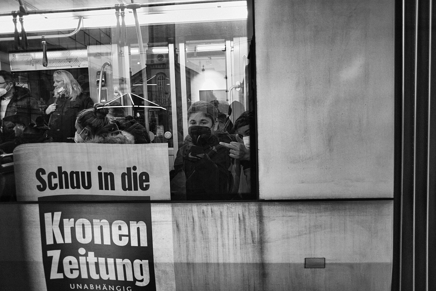 Menschen, die in einem Bus oder Zug sitzen, tragen Masken. Es gibt eine Tafel mit deutschem Text, der die Zeitung 'Kronen Zeitung' bewirbt.