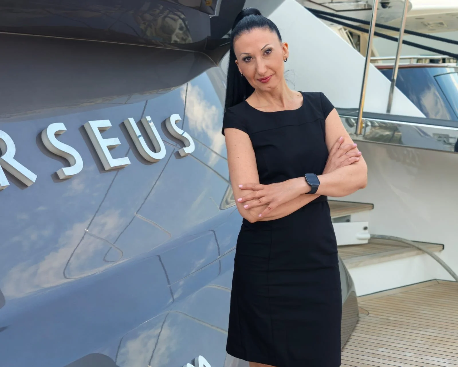 A woman with dark hair tied in a high ponytail, wearing a black dress and smartwatch, standing with her arms crossed in front of a large yacht with the word 'RESOURCES' on its side.