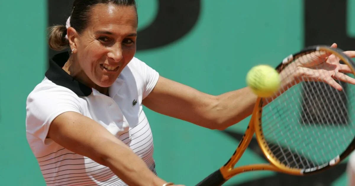 A woman playing tennis, about to hit a yellow tennis ball with a racket, on a green indoor court.