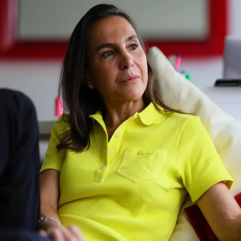 A woman with dark hair wearing a bright yellow polo shirt, sitting in a room with a red-framed whiteboard or window behind her, looking to her left with a serious expression.