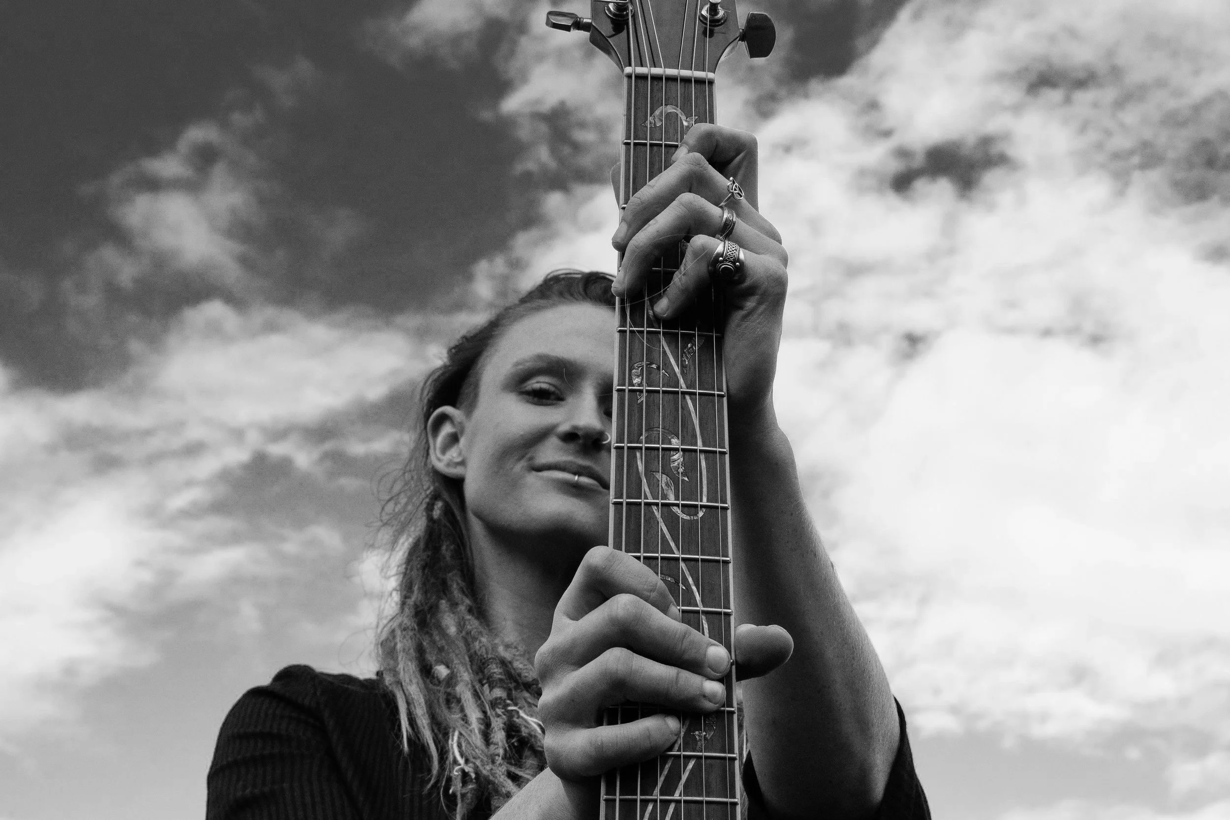 A woman with long hair holding an acoustic guitar up toward the sky, with clouds in the background.