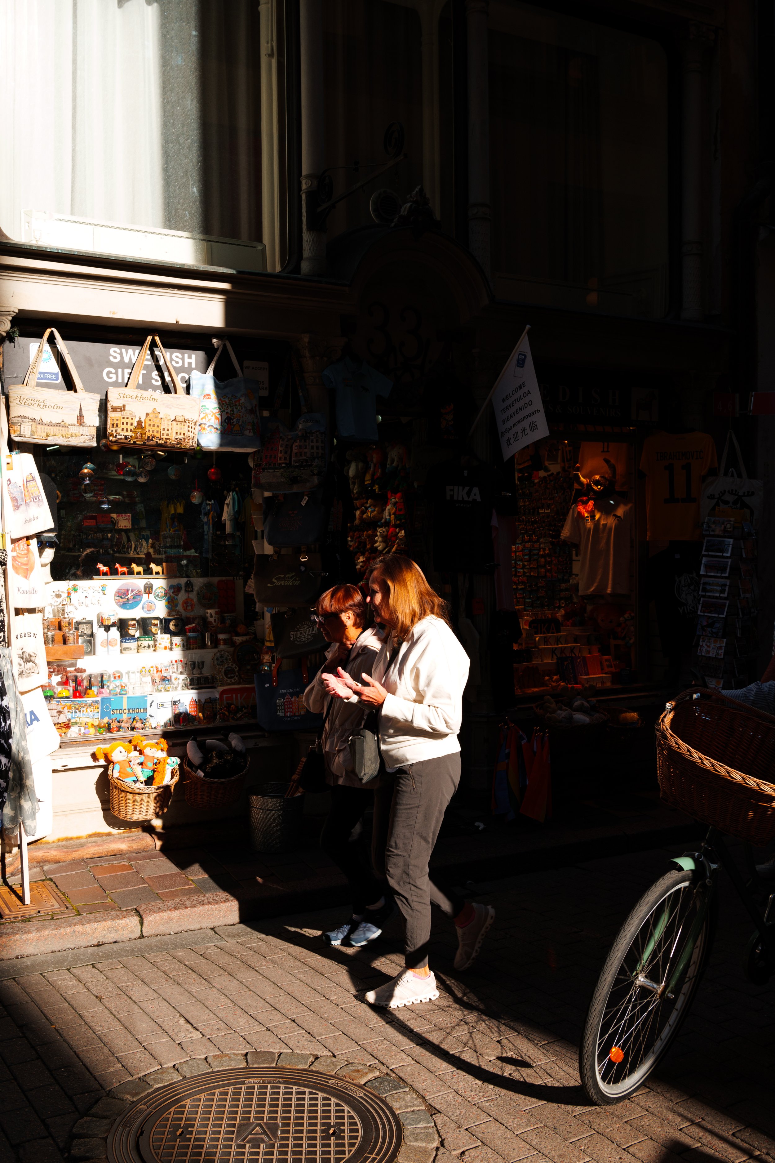 Two women walking past a souvenir shop on a city street, with various items displayed outside, including tote bags and miniature souvenirs.