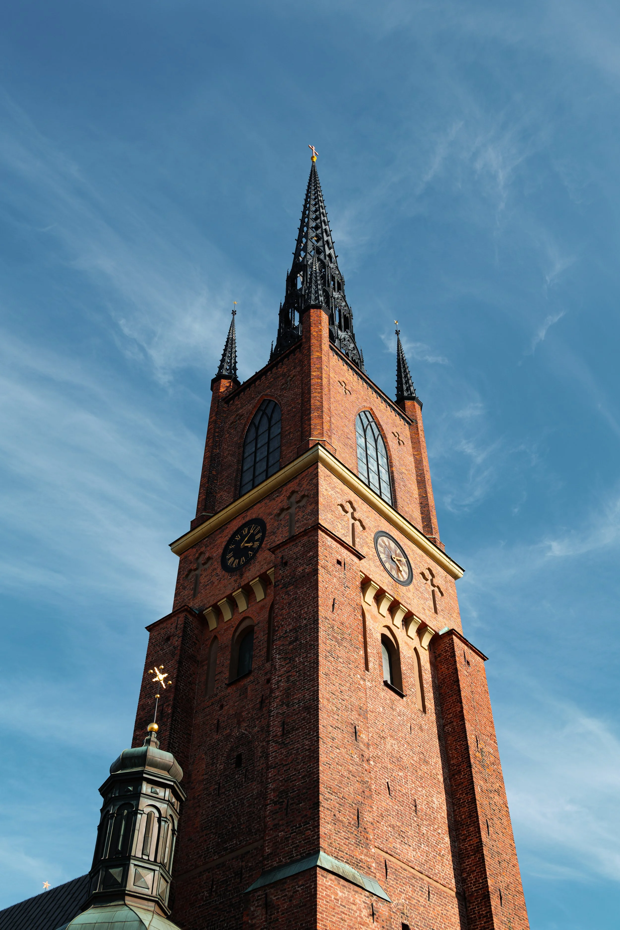 A tall brick church steeple with black spires and clock faces, set against a blue sky with wispy clouds.