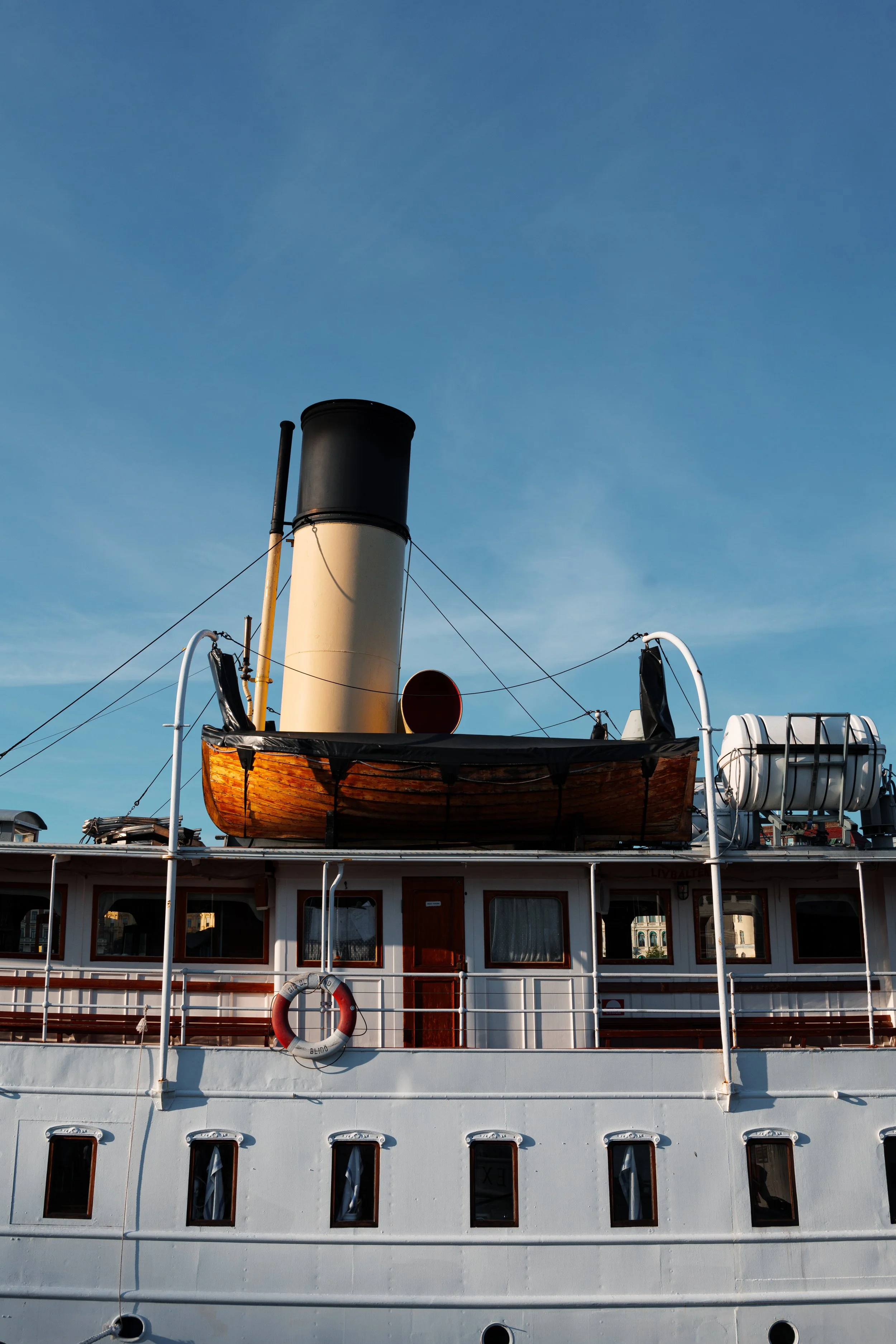A historic passenger steamboat with a large black and beige smoke stack, a wooden boat on the deck, and a life preserver hanging on the railing, under a clear blue sky.