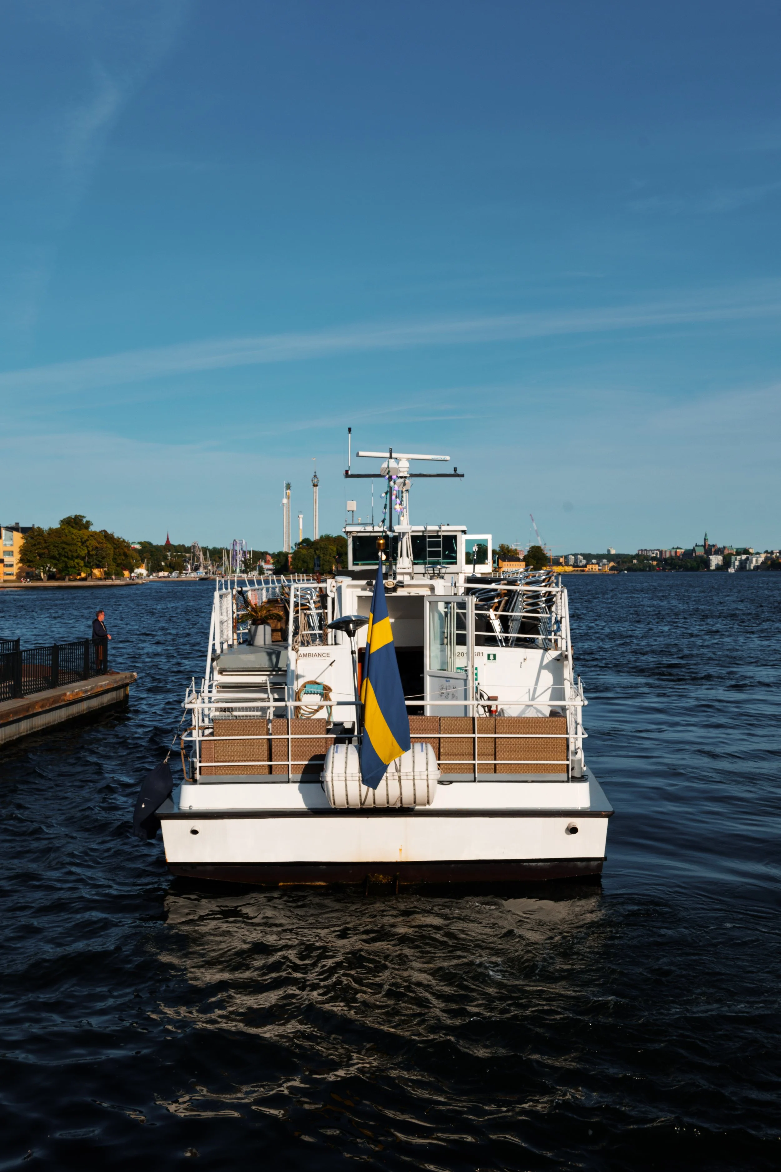 A white boat docked at a pier on a calm river under a clear blue sky, with city buildings visible in the background, and Swedish flag flying at the stern.