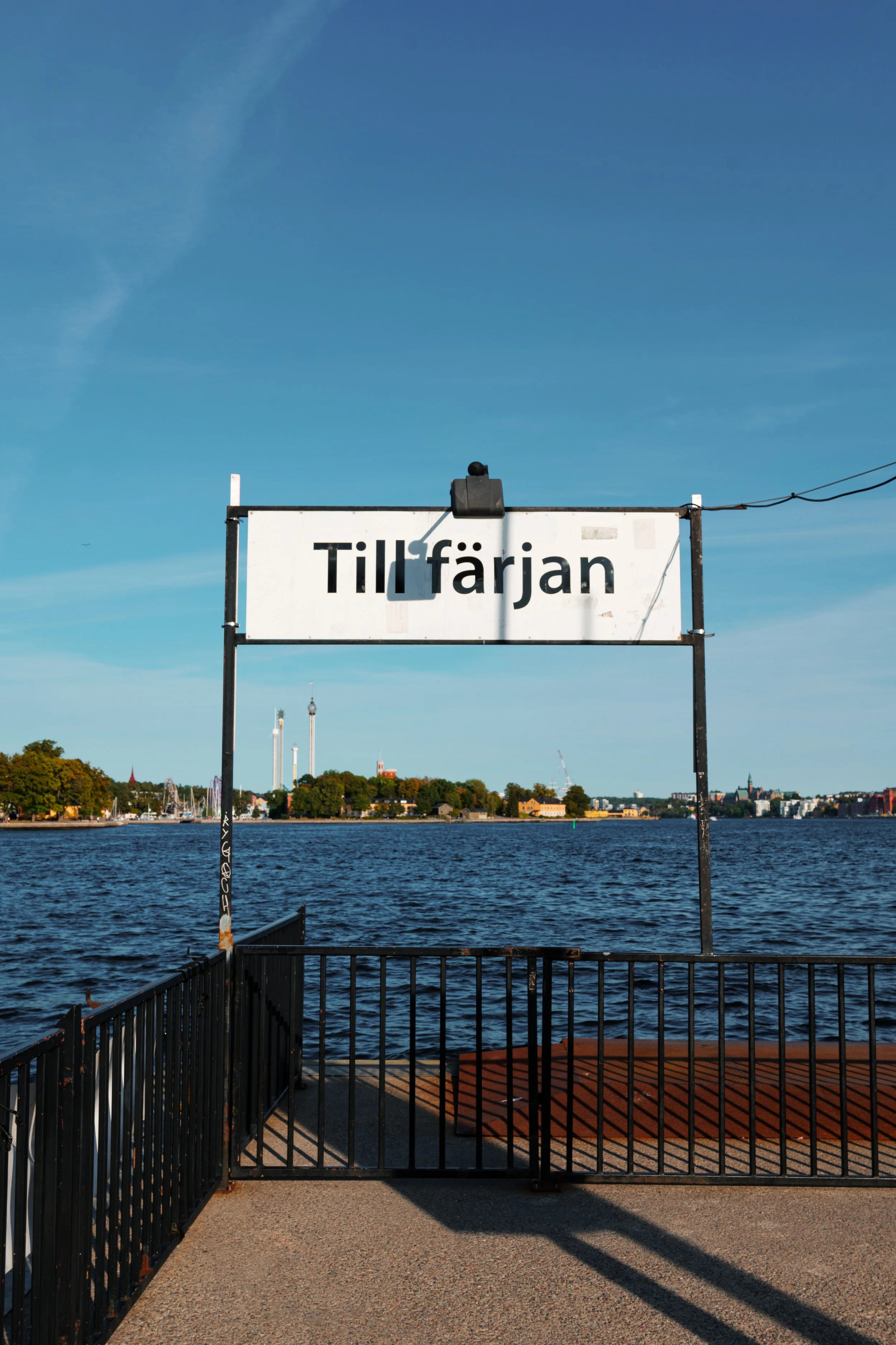 Sign with the text 'Tillfärjan' over water, with distant trees, buildings, sailboats, and a tall chimney in the background under a blue sky.