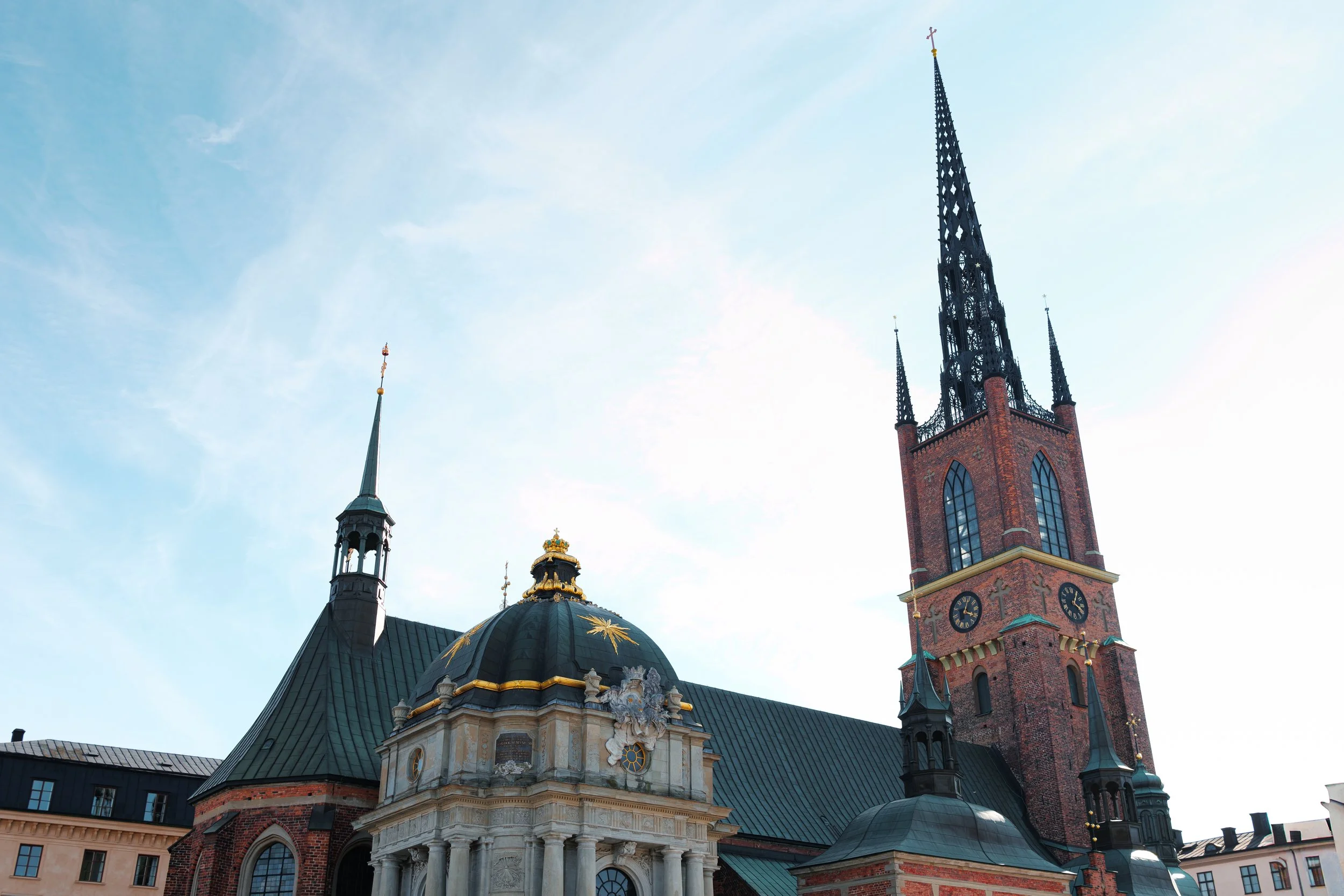 A historic church with red brick and green roofs, featuring tall spires and ornate architecture, set against a clear blue sky.