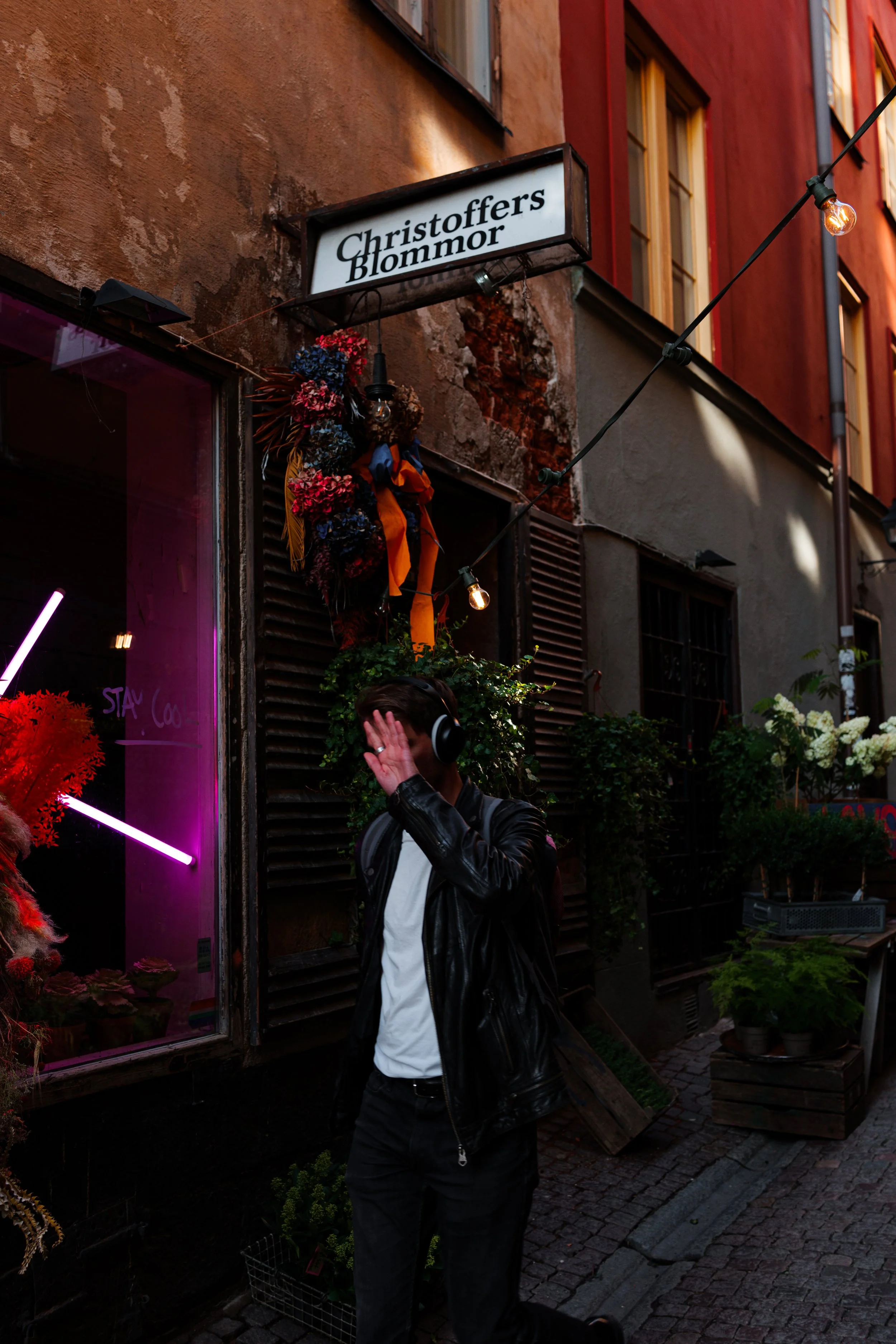 A person wearing a leather jacket and headphones stands on a cobblestone street in front of a store called Christoffer's Blommor, with a colorful floral display and string lights overhead.