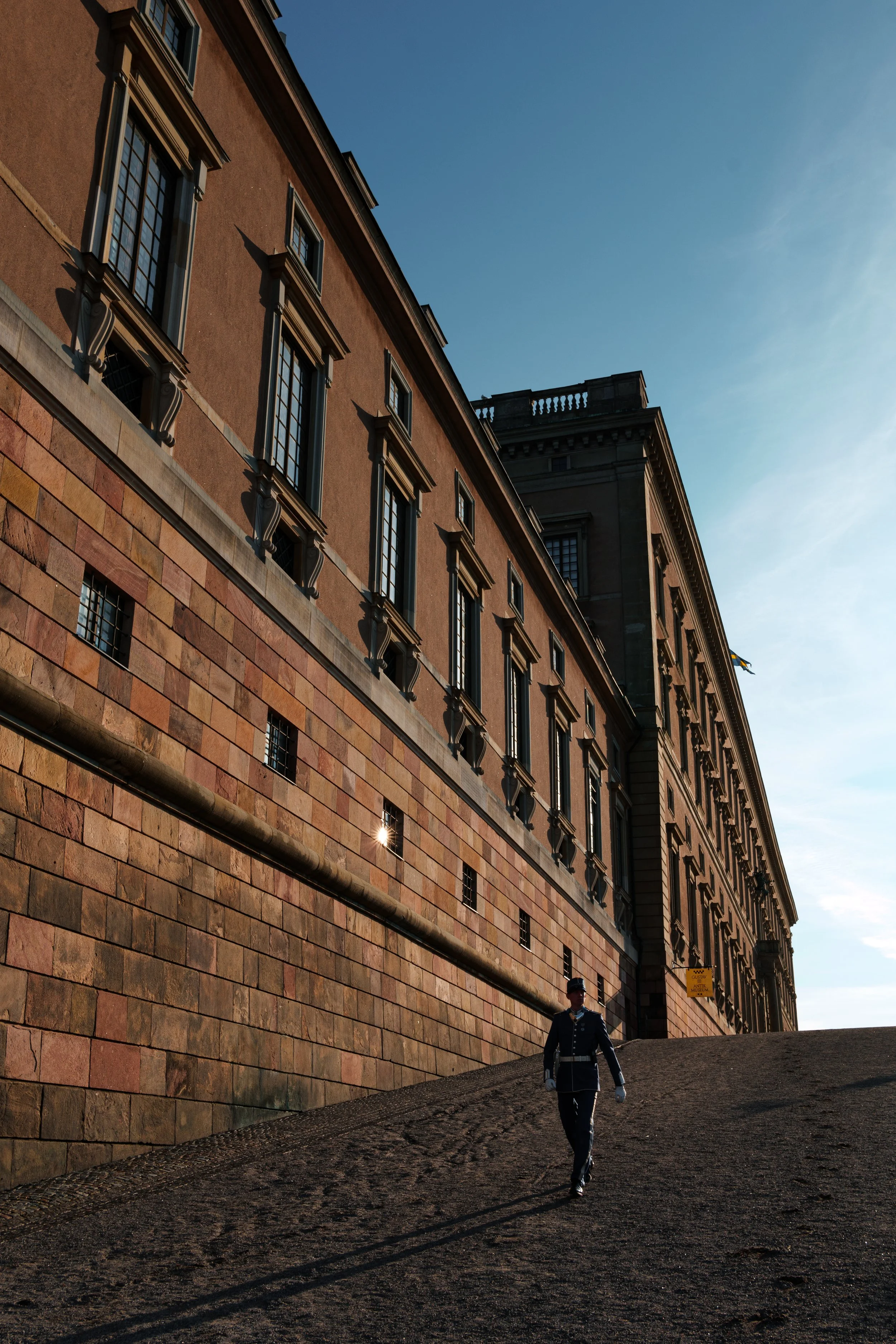 A guard in uniform walking down a dirt path next to a large historic building with multiple windows and detailed architecture, under a clear blue sky.
