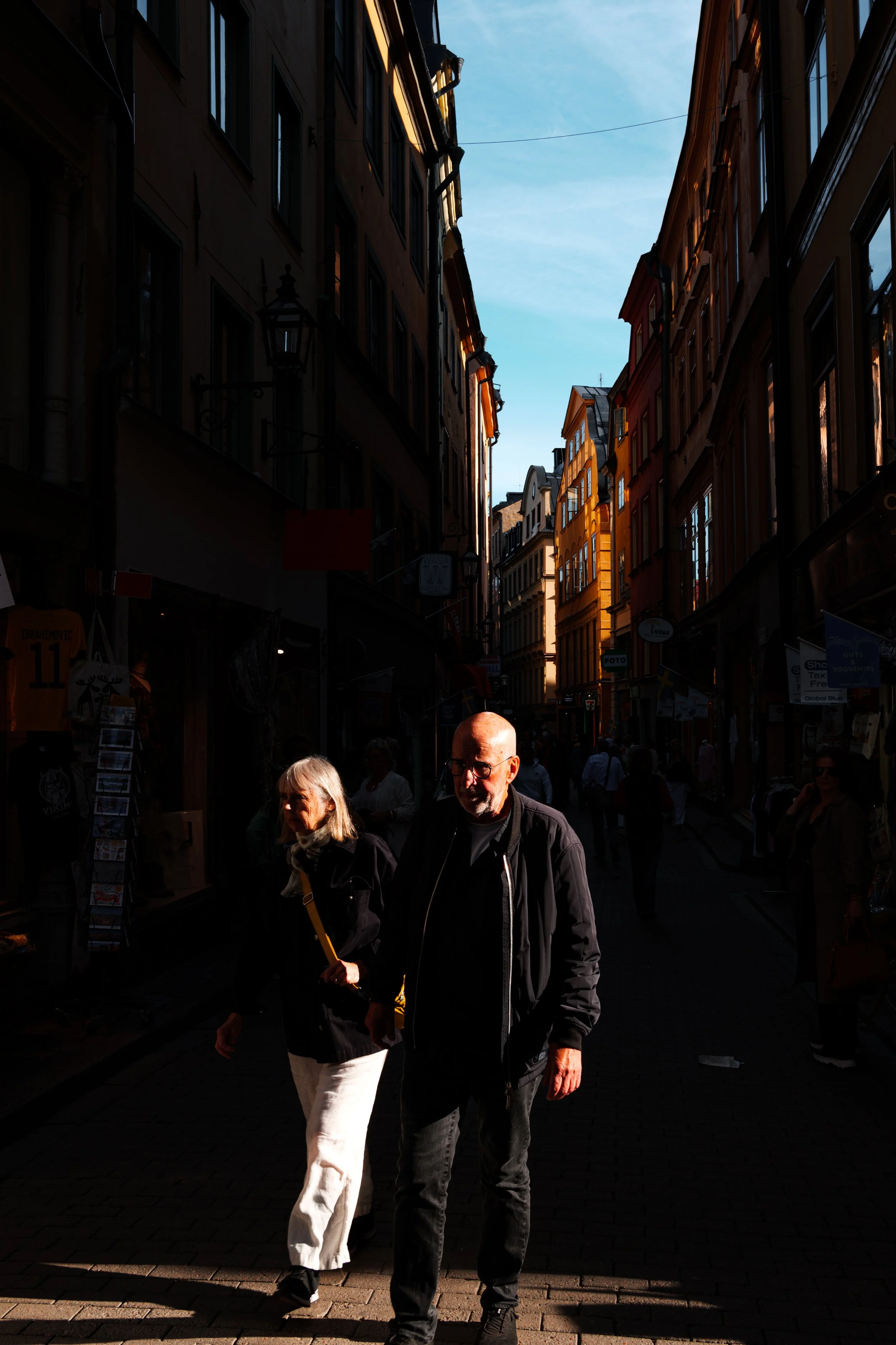 Two people walking down a narrow city street with tall buildings on either side, illuminated by sunlight with the sky visible above.