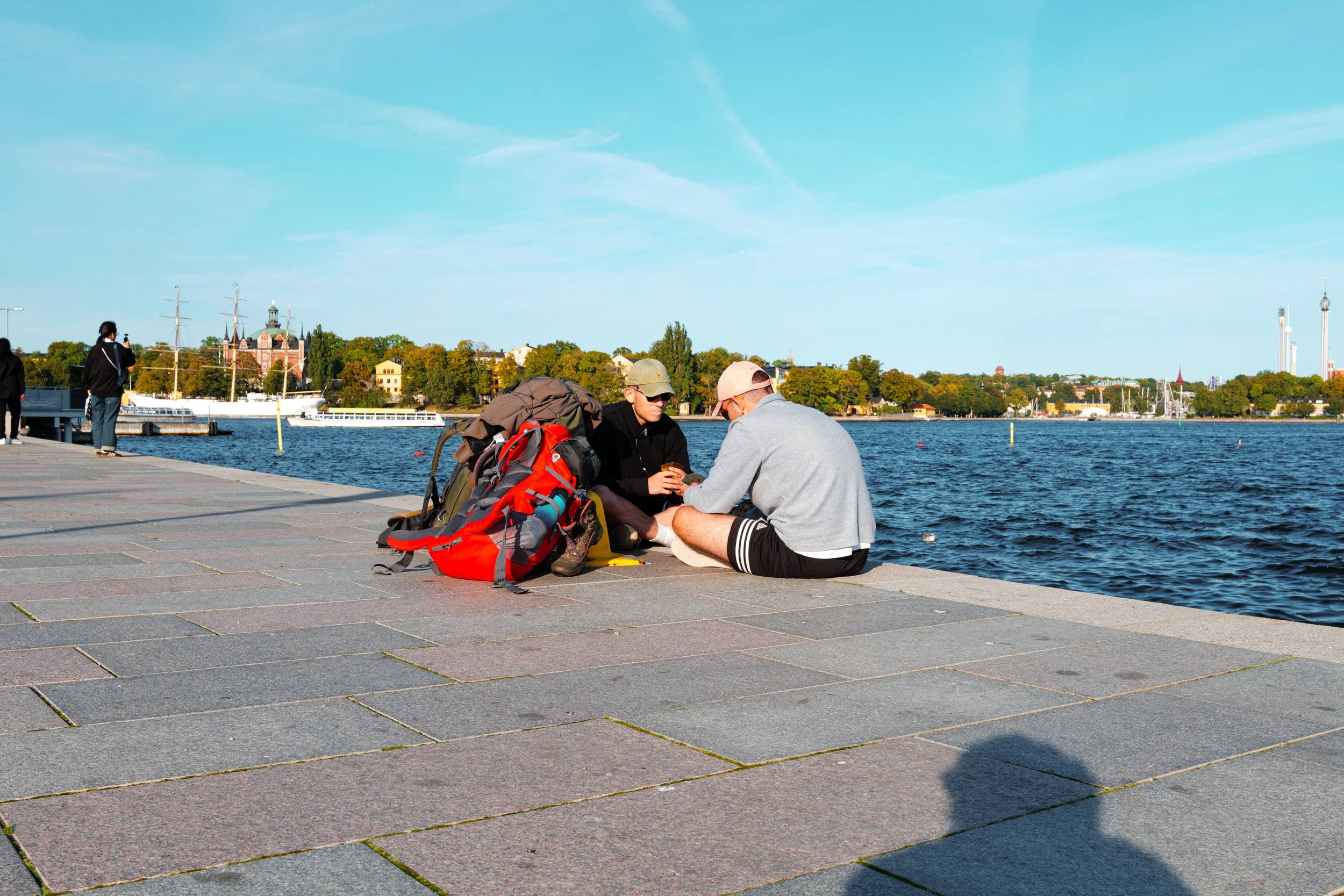 Two young men with backpacks sitting on the dock by the river, having a conversation.