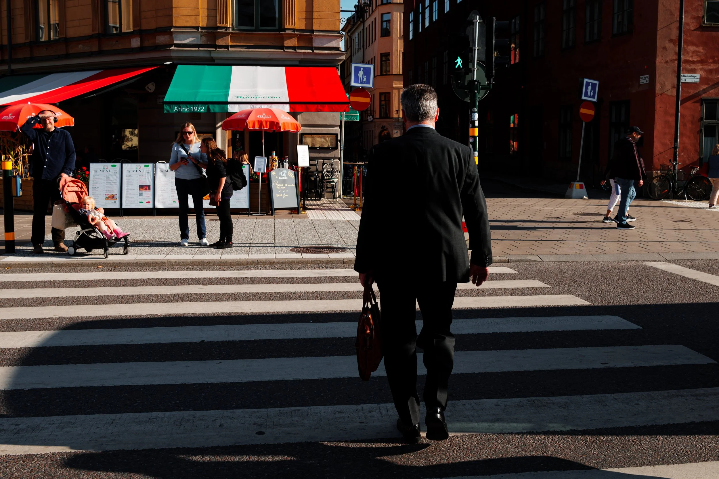 A man in a black suit crossing a city street with a bag in hand, while several pedestrians stand near a cafe with red umbrellas, and others walk along the sidewalk on a sunny day.
