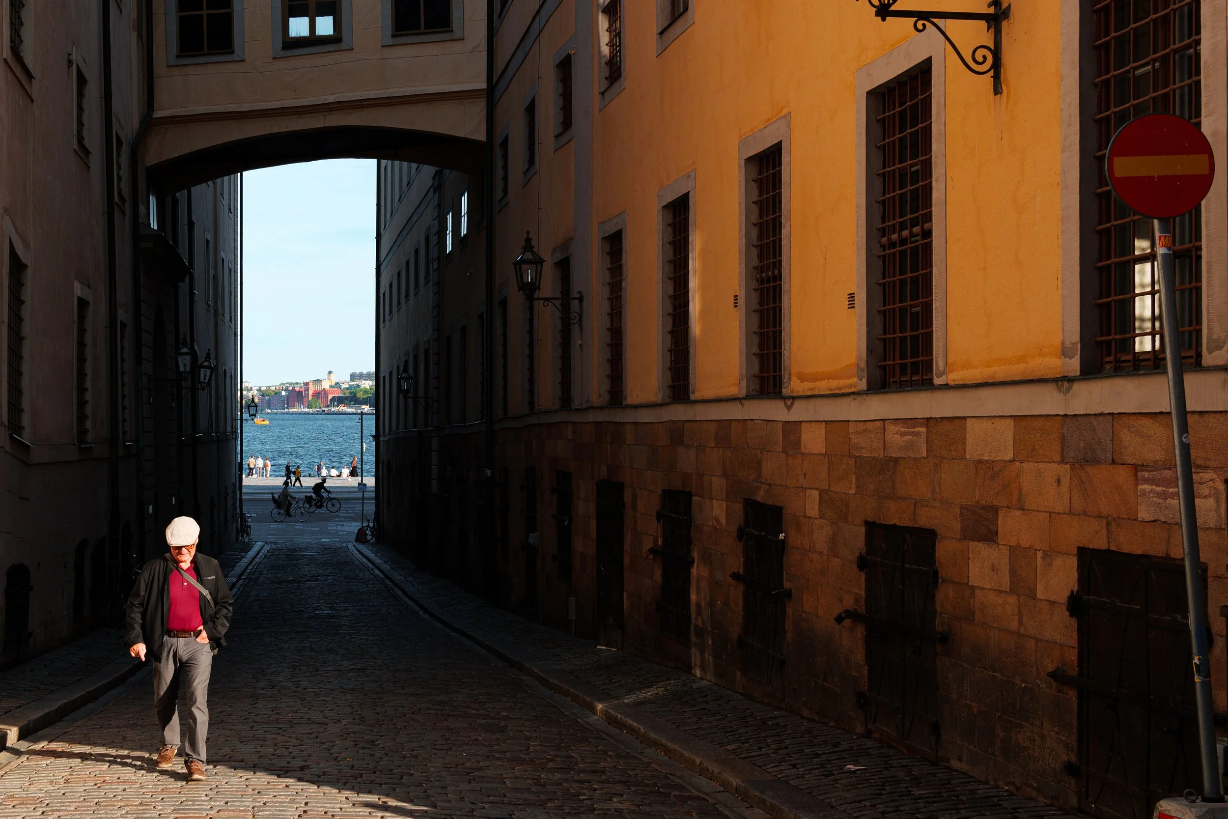 A man walking down a cobblestone alleyway with yellow and beige buildings, leading to a waterfront with people and bicycles, and a view of water and buildings in the distance.