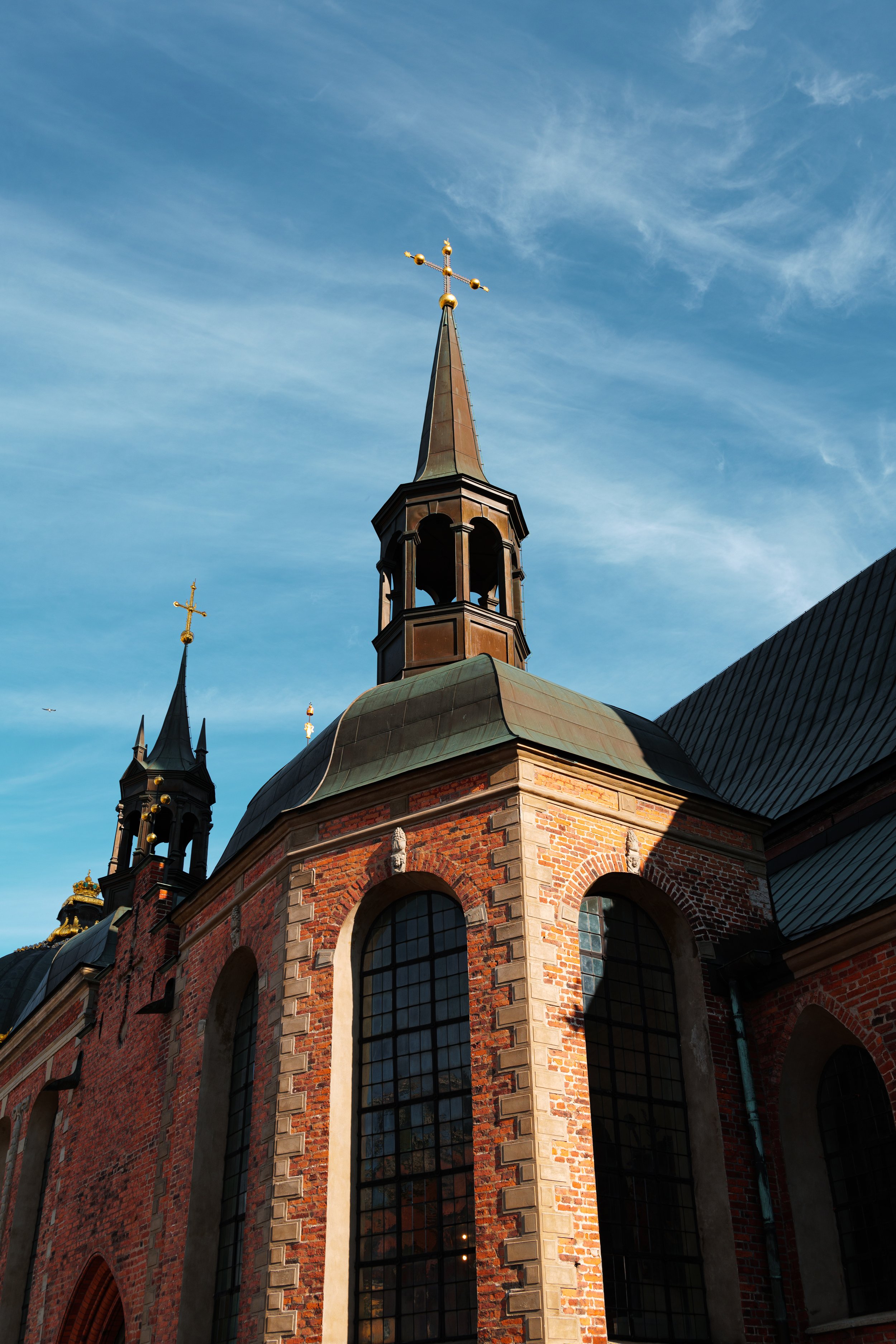 A historic brick church with tall windows and steeples topped with cross symbols, set against a bright blue sky with wispy clouds.